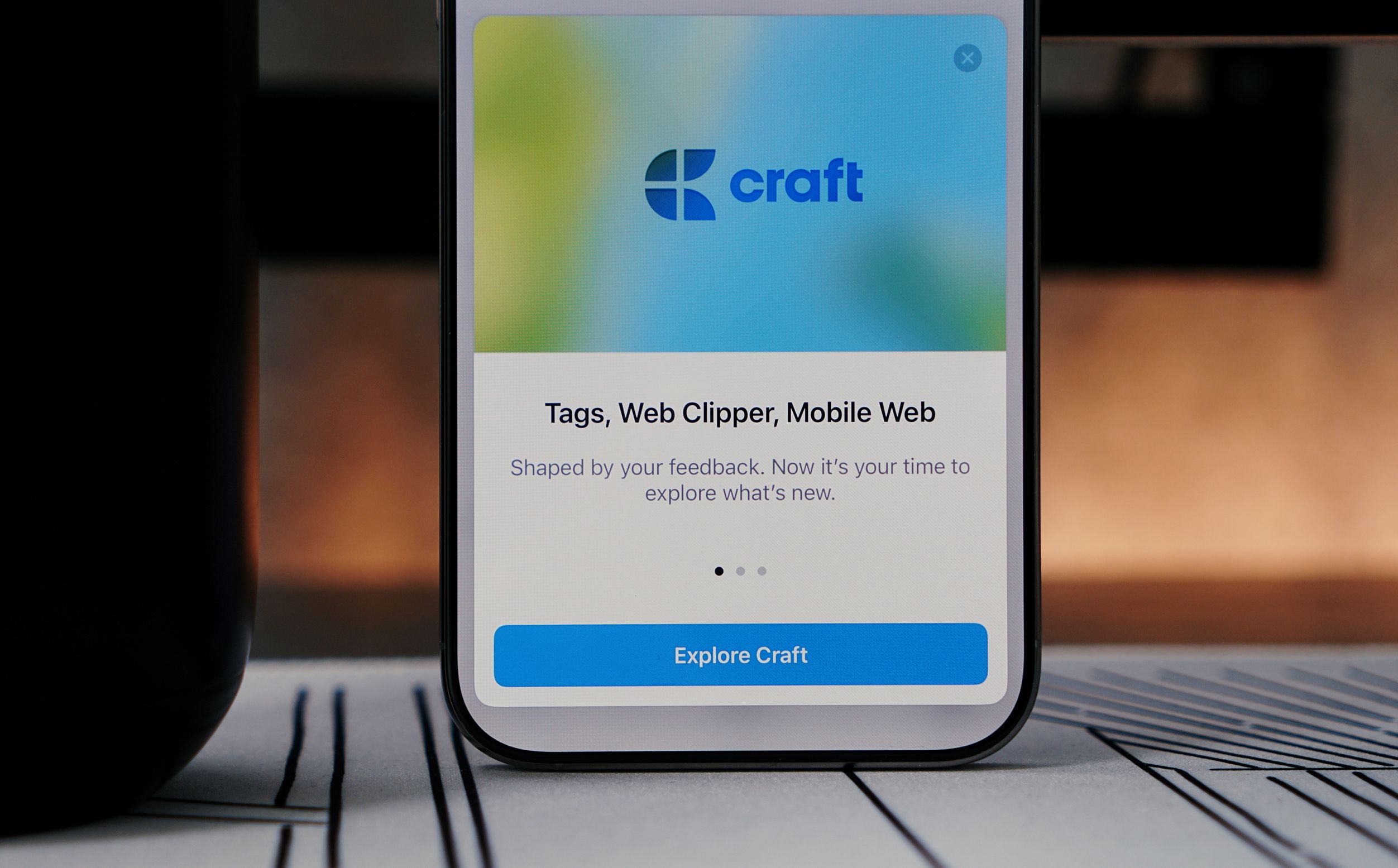 A smartphone displaying the Craft app welcome screen. The screen has a blurred blue and green background with the Craft logo at the top, and the text "Tags, Web Clipper, Mobile Web" followed by a description and an "Explore Craft" button. The phone sits on a patterned surface next to a black object, likely a cup.