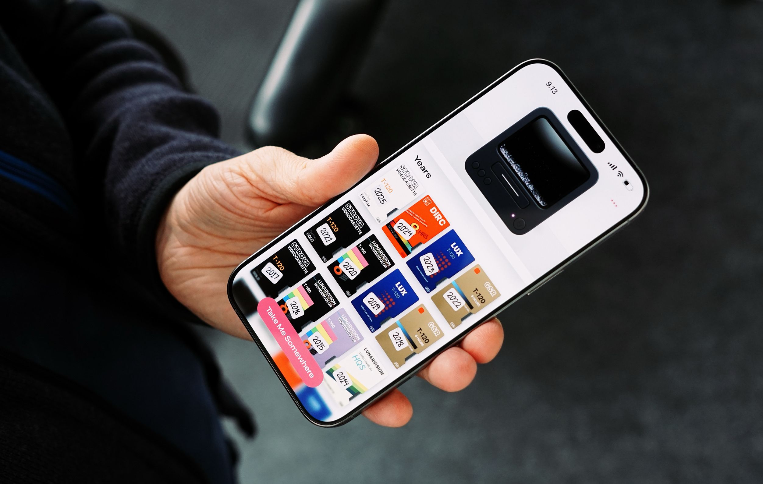 A hand holding an iPhone displaying a user interface with the title "Years" and various blocks representing different years, each block featuring text related to video cassette tapes, the interface includes a "Take Me Somewhere" call to action.
