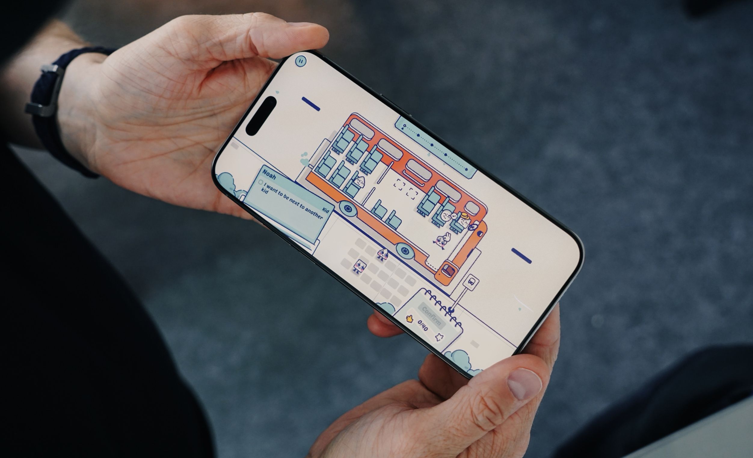 A smartphone held by hands displays a cartoon bus interior with characters, likely a mobile game. The screen highlights a seating arrangement interface with dialogue bubble "I want to be next to another kid" along with confirmation and currency options.
