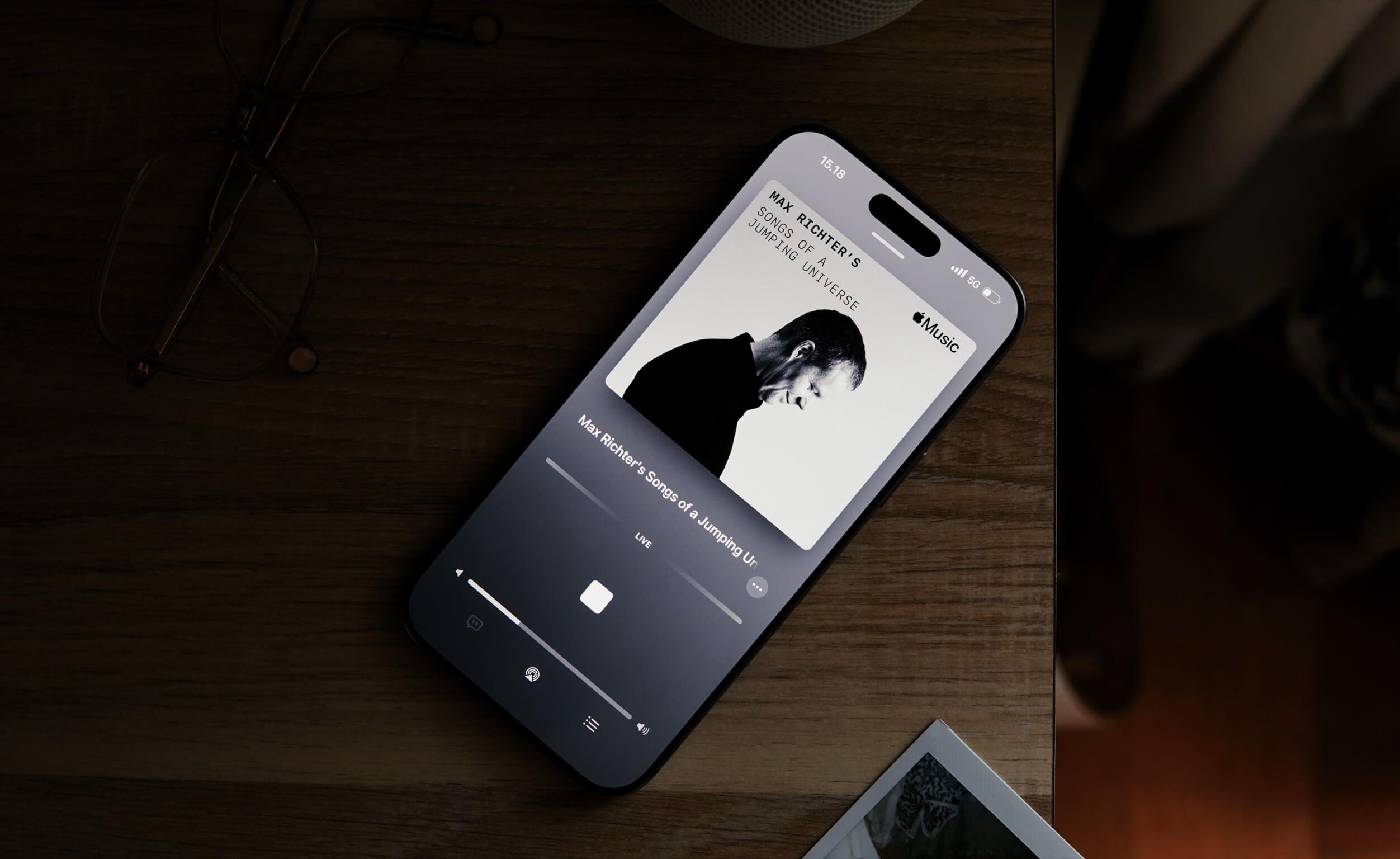 A smartphone displaying the Apple Music app with Max Richter's "Songs of a Jumping Universe" playing, lying on a wooden surface with glasses on top.
