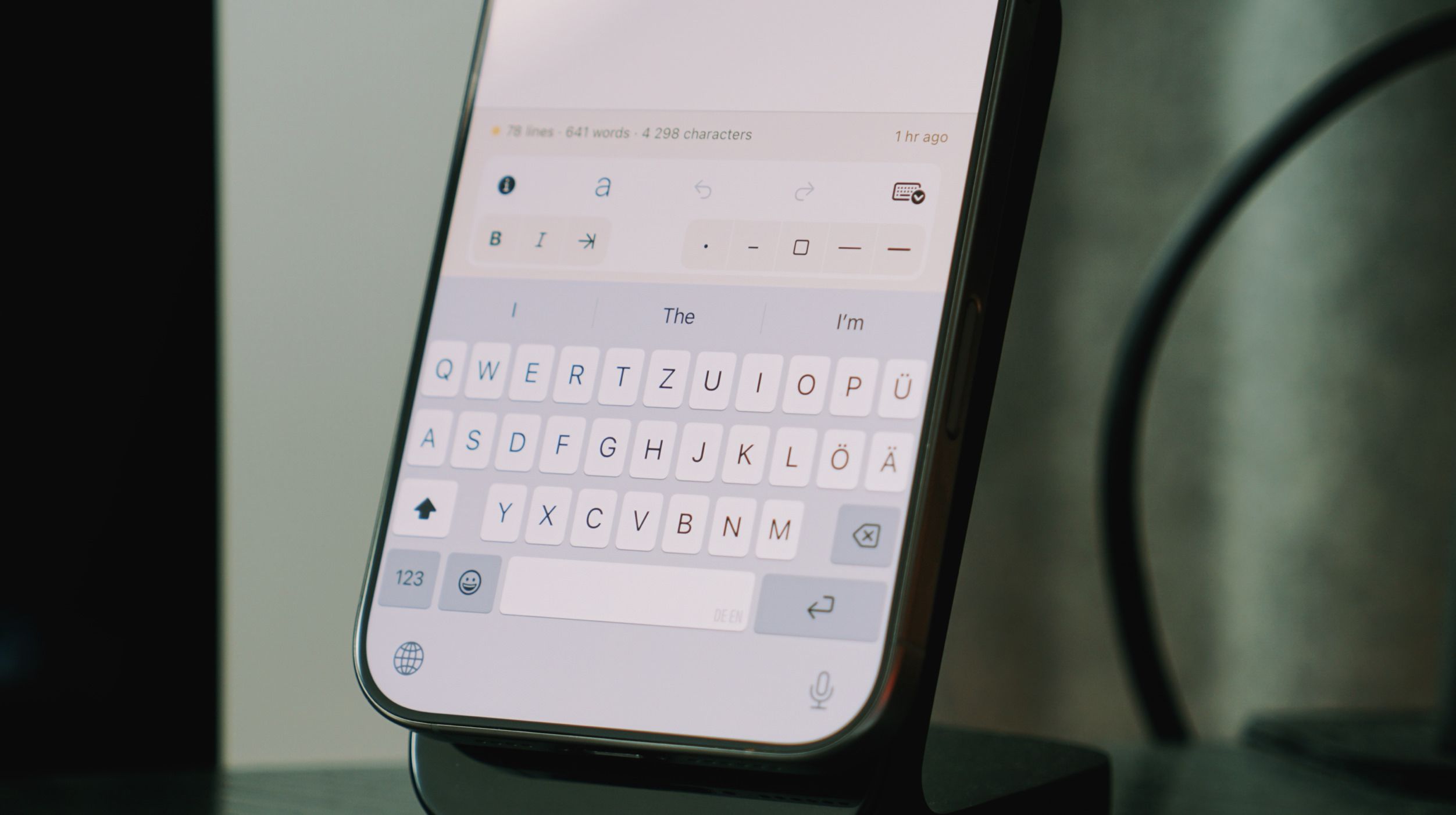 Close-up of a smartphone screen displaying a writing interface with a keyboard, likely a document, resting on a black stand. The screen's interface shows text formatting options above the keyboard, with the letters "QWERTY..." visible. The screen displays the words "The" and "I'm" as context for the text.
