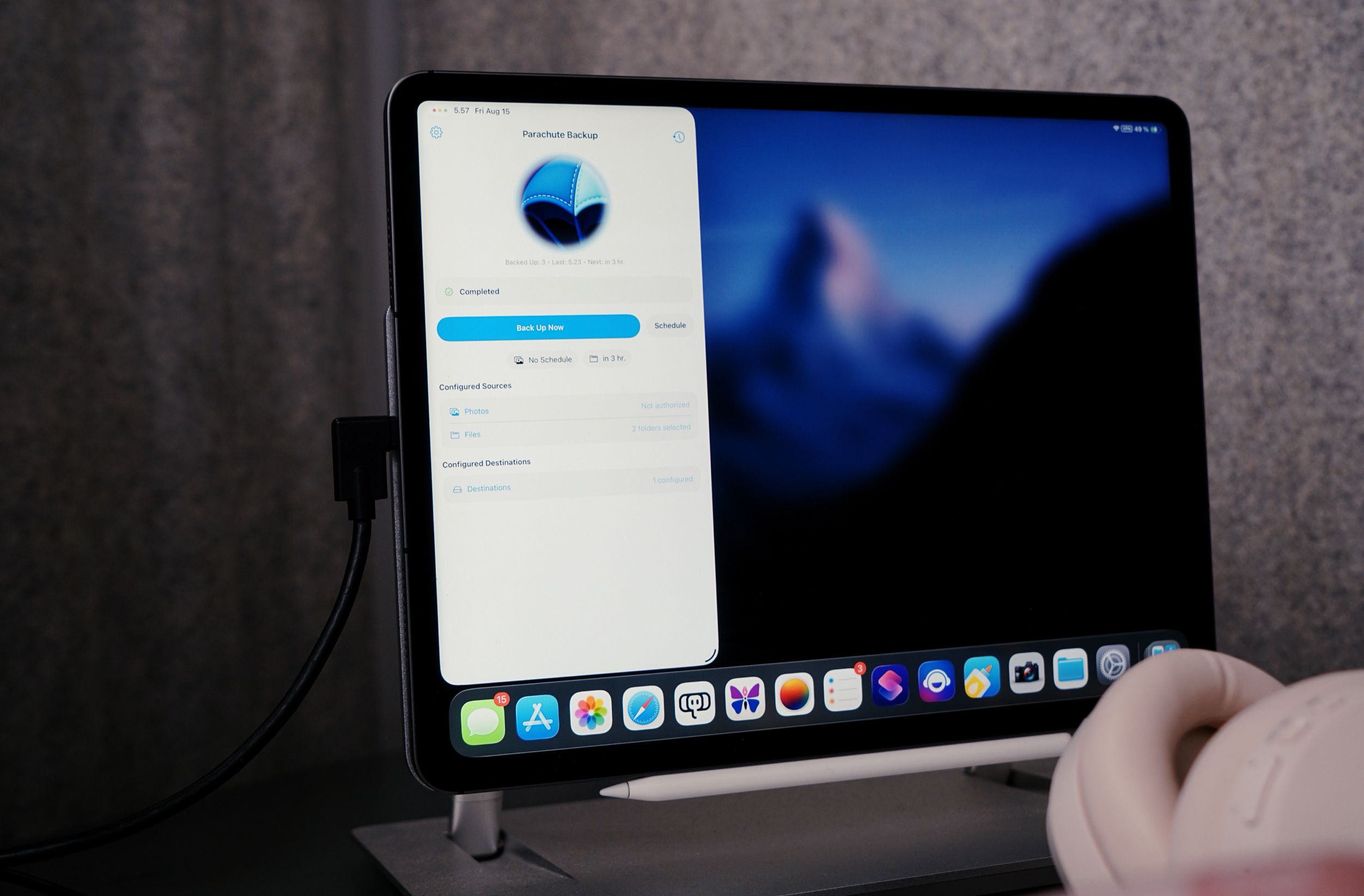A close-up of an iPad displaying the "Parachute Backup" app with a progress overview. The screen shows the backup status as "completed," with options to "Back Up Now" or "Schedule". The app details the backup configuration, including sources like "Photos" (not authorized) and "Files", and destinations. The iPad is connected to a power cord and is placed on a stand. On the bottom of the screen, there are several app icons, and there's also a white pencil. In the lower right corner are white headphones partially visible.
