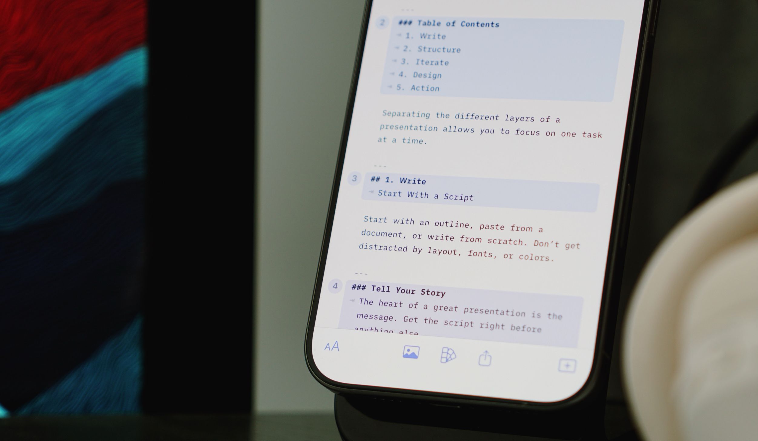 Close-up shot of a smartphone displaying a presentation outline with numbered sections on writing a presentation, with a table of contents including Write, Structure, Iterate, Design, and Action. The screen shows content related to scripting a presentation. The phone is set against a blurred background with abstract red and blue color patterns and a white cup on the right.
