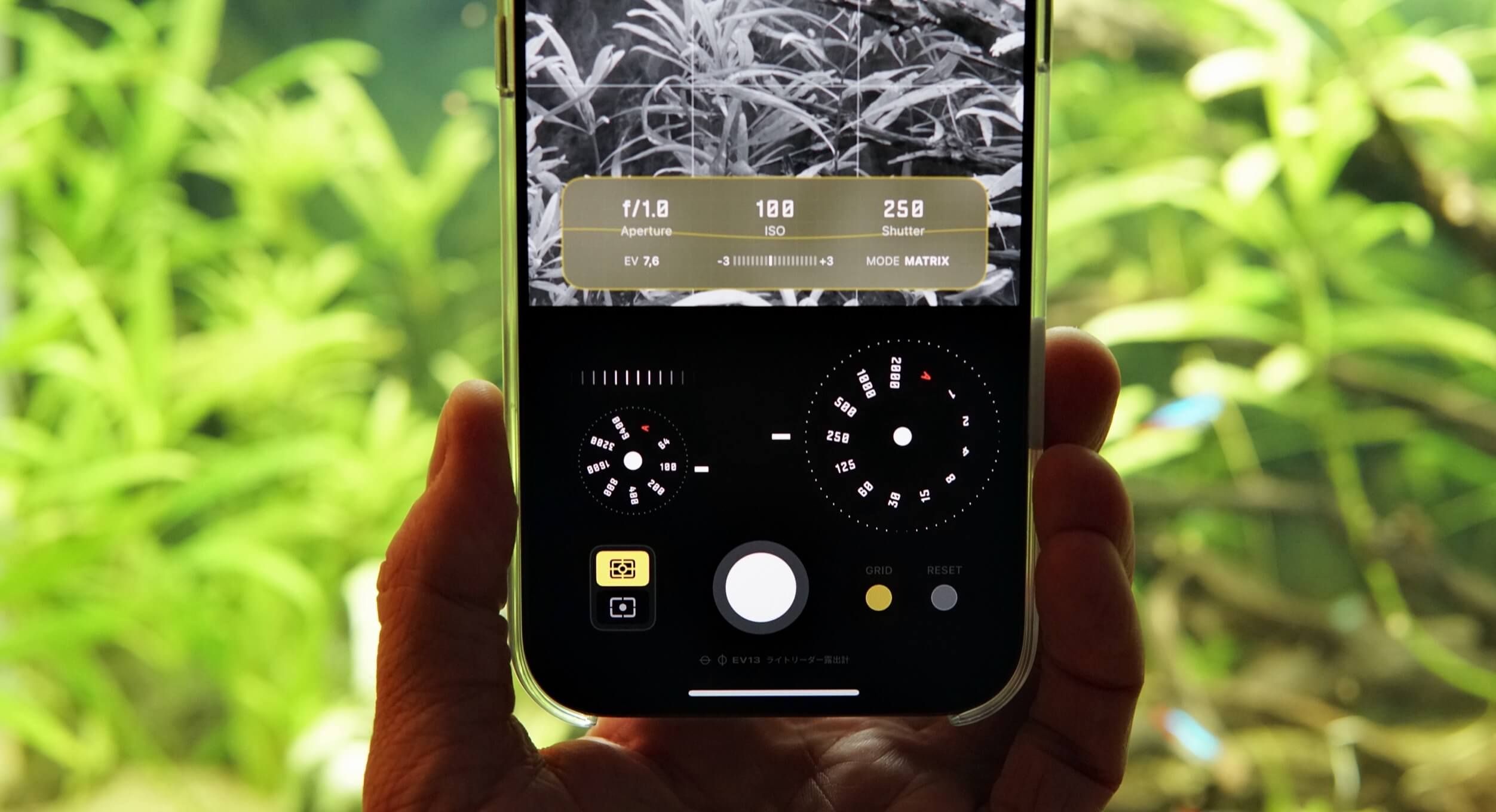 Hand holding a smartphone displaying a camera app interface with black and white preview of greenery, showing aperture, ISO, and shutter speed settings at the top, a settings dial, and other controls; background is blurred green foliage.
