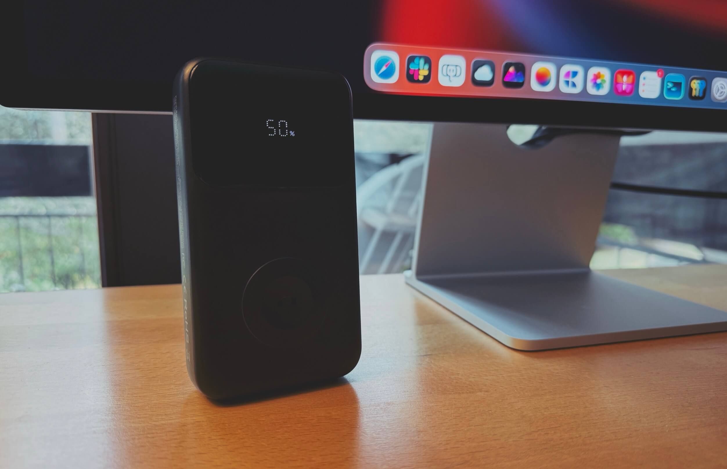 A black portable charger displaying "50%" on a digital screen, placed on a wooden desk with a computer monitor and stand in the background.