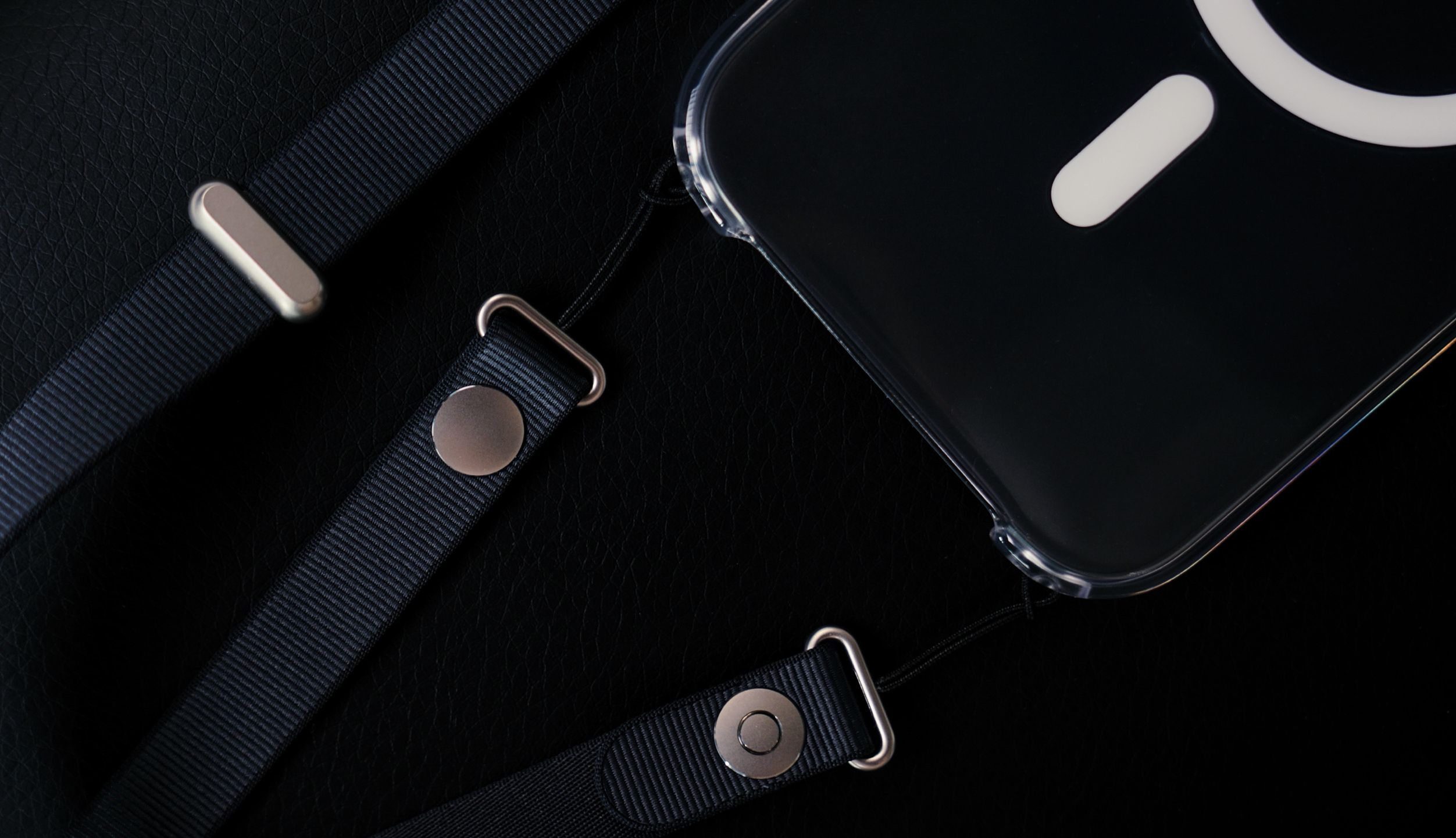 Close-up image of a clear phone case with an attached lanyard and metallic hardware, resting on a textured black surface. The case has a white, circle and rectangle design. The lanyard is made of dark blue, woven fabric, attached with silver colored metal loops, snaps, and bars.
