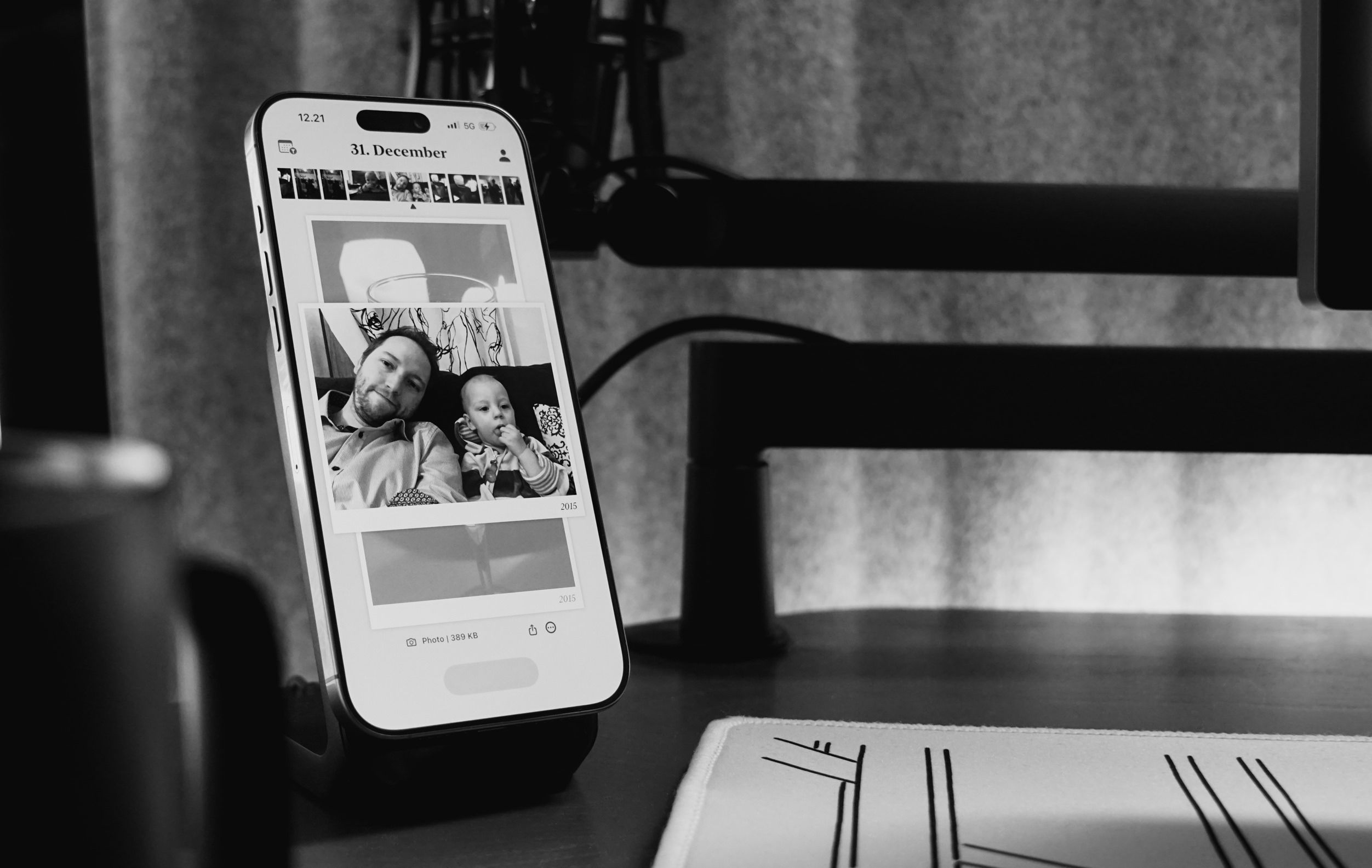 Black and white image of a smartphone displaying photos, held in a charging dock on a desk. The phone shows pictures of a man and a baby, taken on December 31st, 2015. A microphone and desk components are visible in the background, along with a graphic mouse pad.
