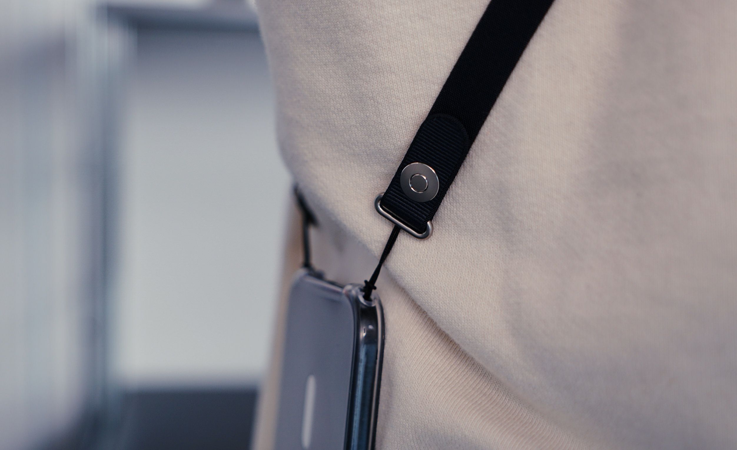 Close-up of a cream-colored hoodie with a black lanyard strap attached. The strap is connected to a clear phone case with a black string, the strap has a metal clasp and a silver button. The background is a blurred, light-colored wall.

