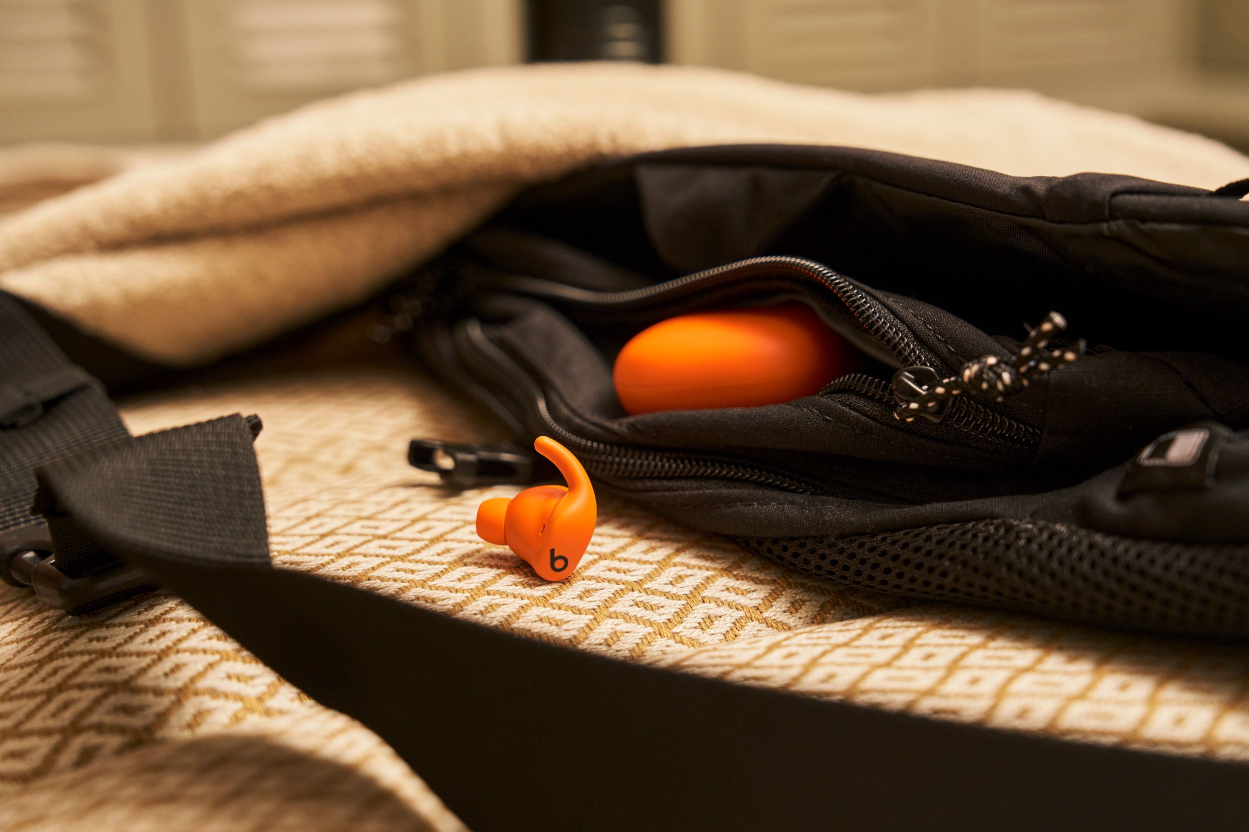 An orange Beats earbud with black "b" logo sits next to a black bag, partially open. The bag reveals a zipped pocket containing another orange item and lies atop a patterned tan surface. A black strap and a fluffy tan fabric are also in the frame.
