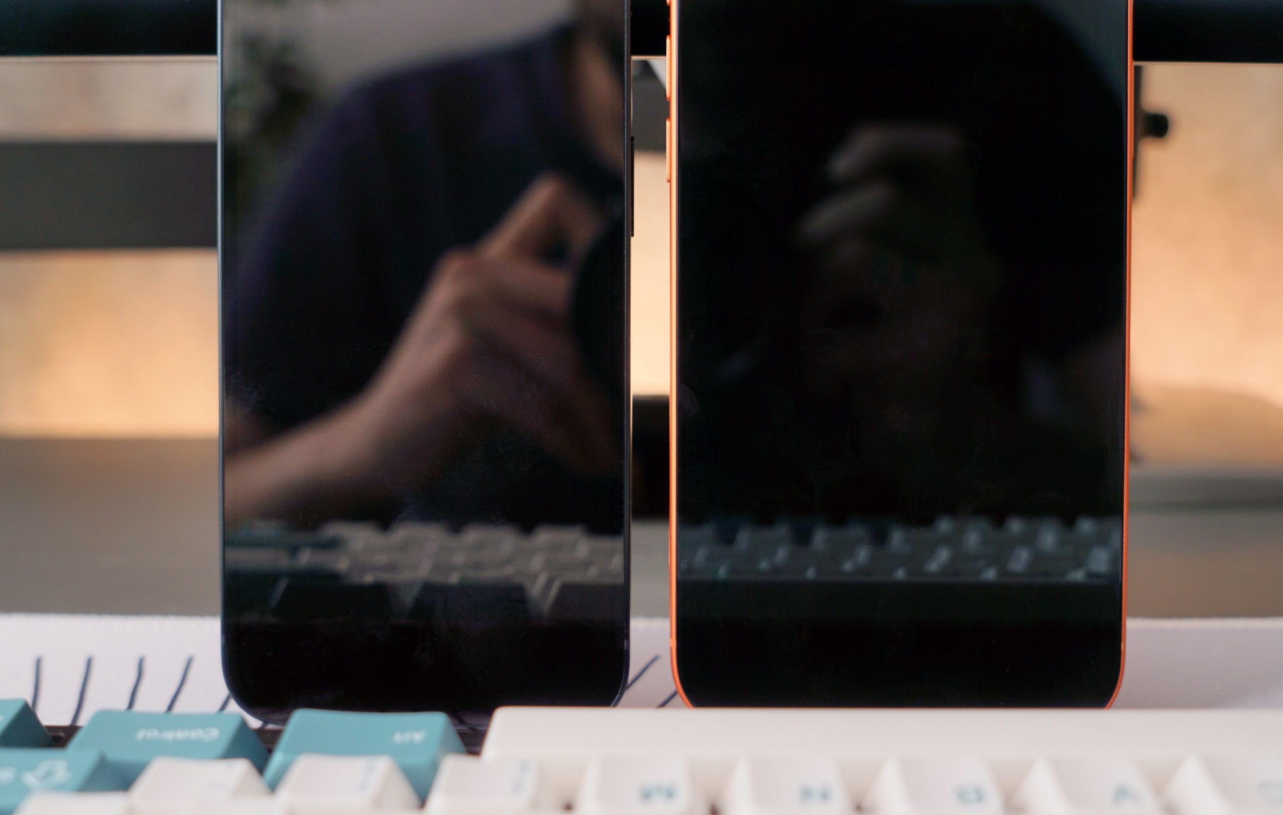 Two smartphones with black screens reflecting a person and keyboard, and an orange-colored frame sitting on top of a white and teal mechanical keyboard.
