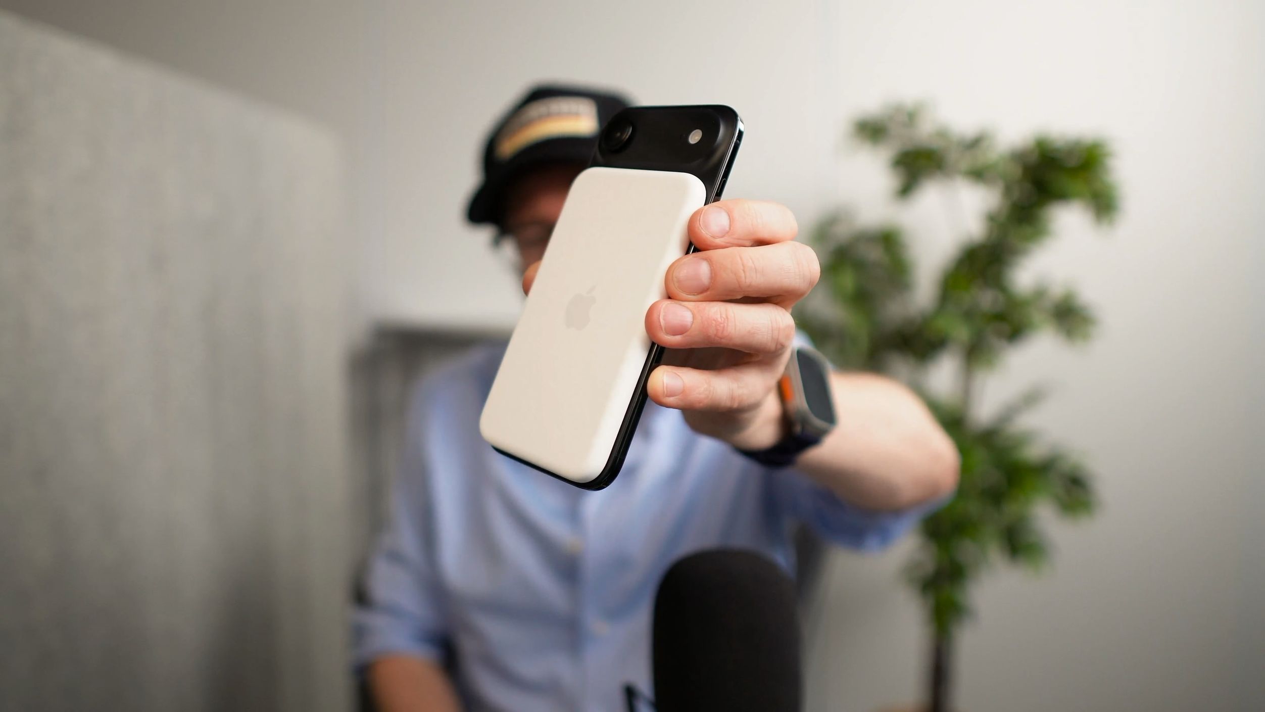 A person, wearing a hat and glasses, holding up a white and black smartphone with the Apple logo visible on its back; background includes a blurred plant and a microphone.

