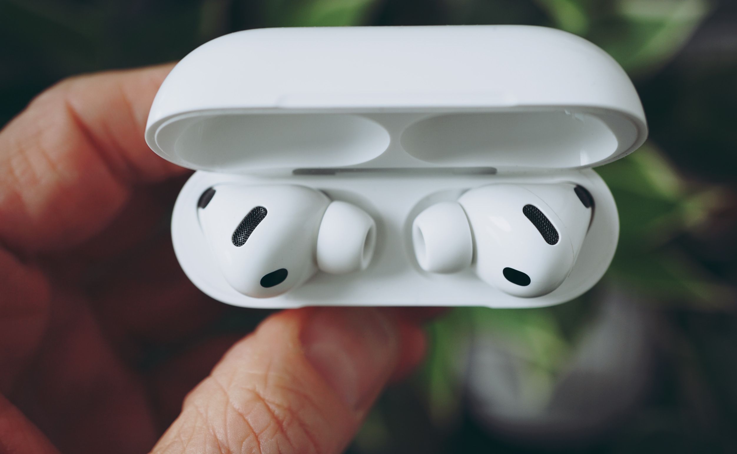 White Apple AirPods Pro earbuds in open charging case, held in a person's hands, with blurred natural background.
