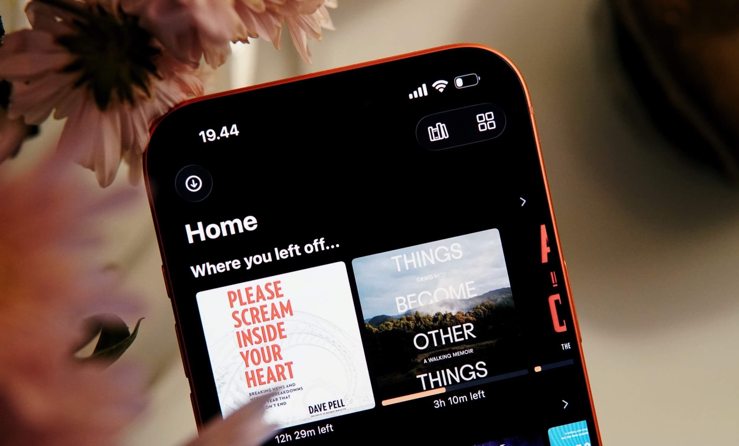 Close-up of a smartphone screen displaying a home screen for an audiobook app, with a dark interface featuring two book covers: "Please Scream Inside Your Heart" and "Things Become Other". The phone is red with a black screen and a flower in the background.
