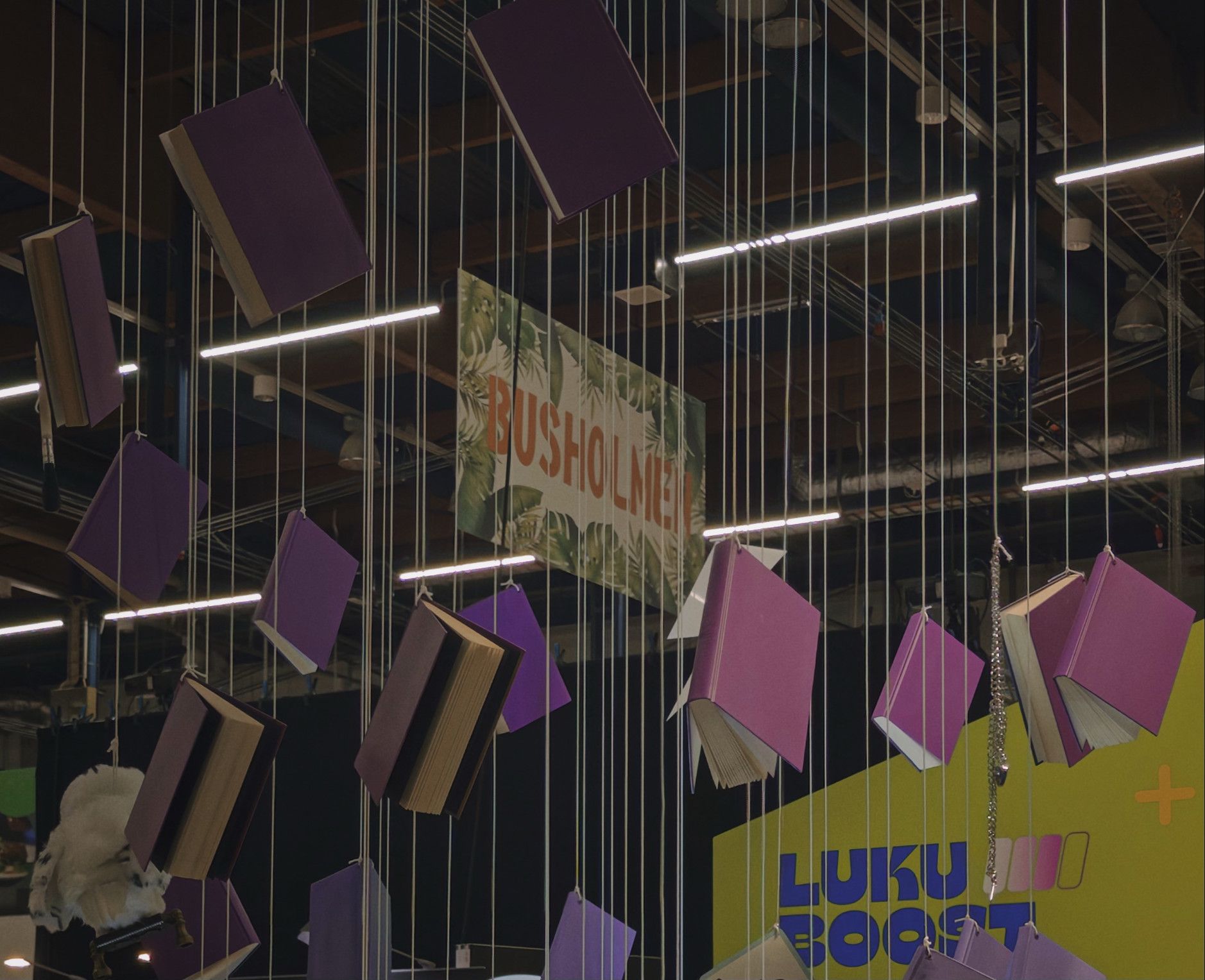 Books with purple covers, some open, suspended by white strings from the ceiling, with a sign reading "Busholmen" and a yellow wall with text in the background.
