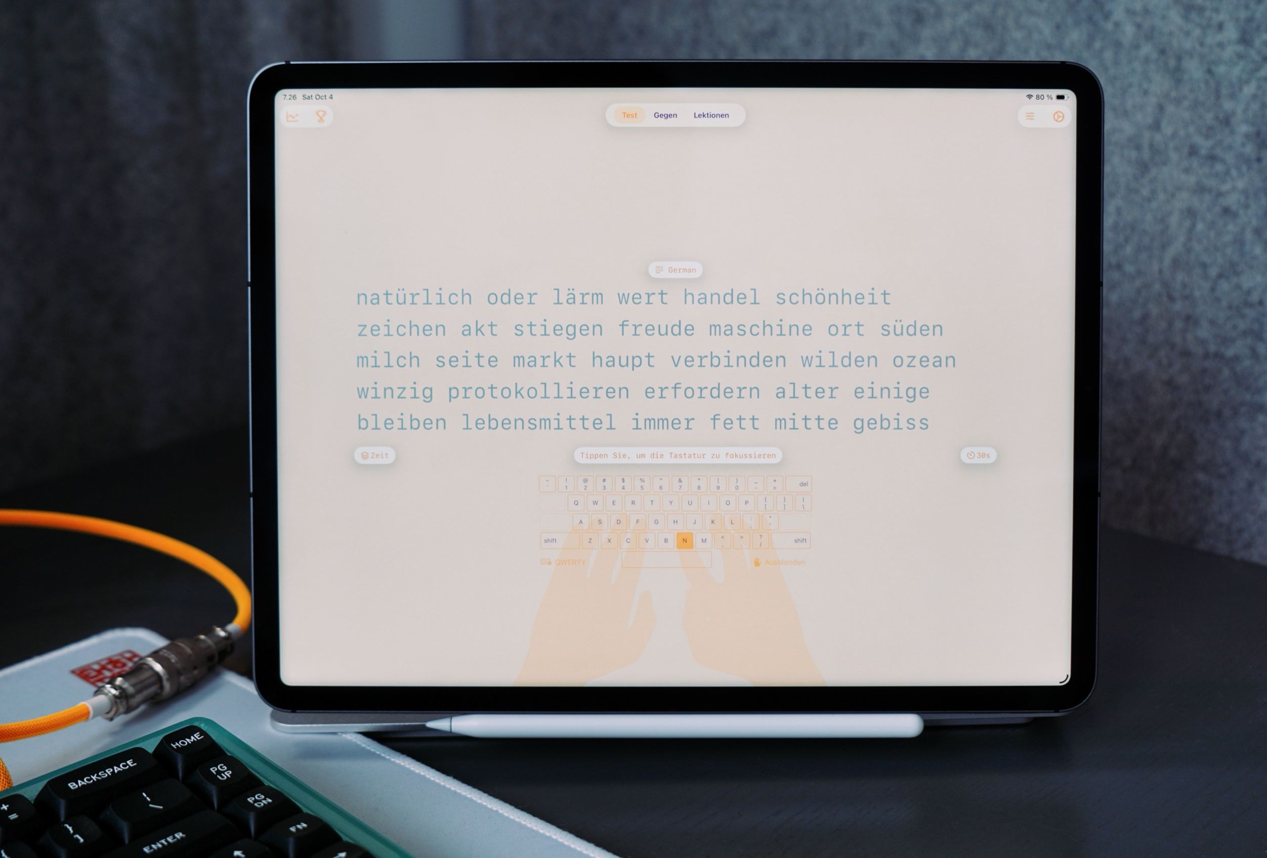 An iPad displays a text-based German lesson on its screen, with a keyboard visible at the bottom, a stylus below the screen, and a mechanical keyboard and cabling in the foreground.