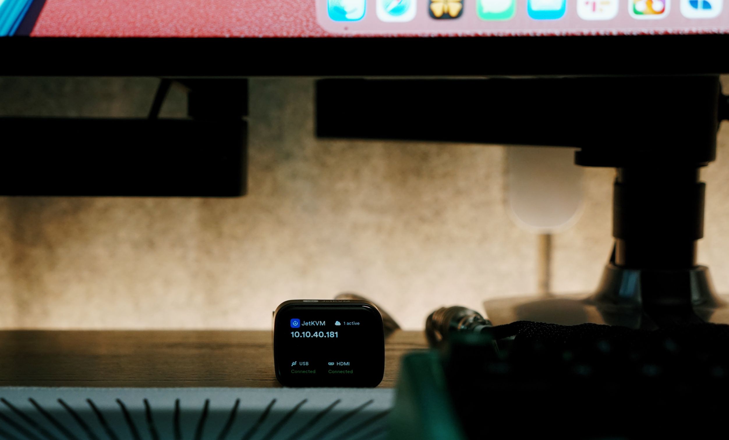 A small, black JetKVM device displays its IP address, 10.10.40.181, with USB and HDMI connections indicated as connected, resting on a dark surface in front of a computer monitor, partially obscuring the monitor's display.