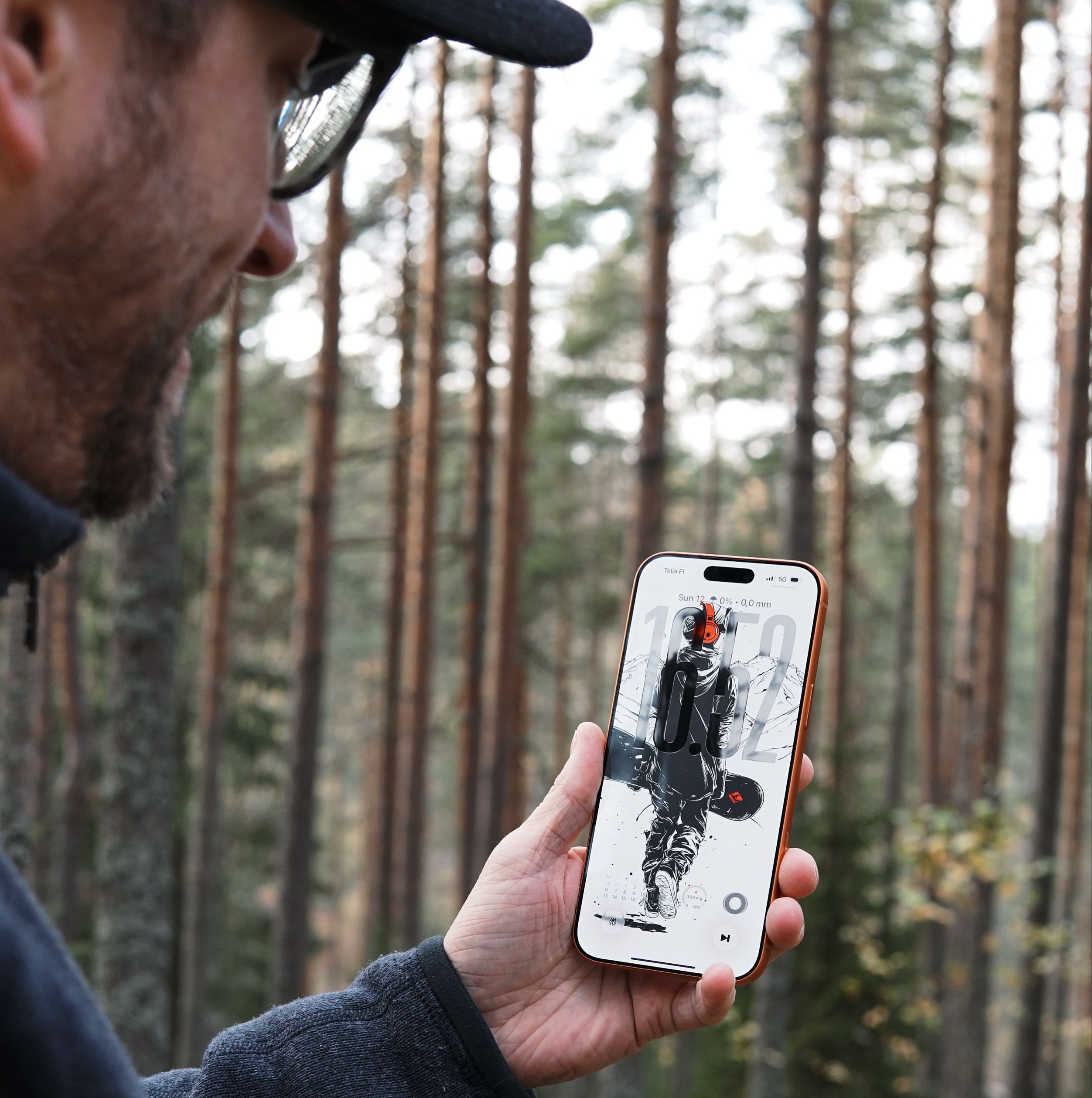 Man holding a smartphone with a wallpaper depicting an astronaut with a snowboard, in a forest setting.
