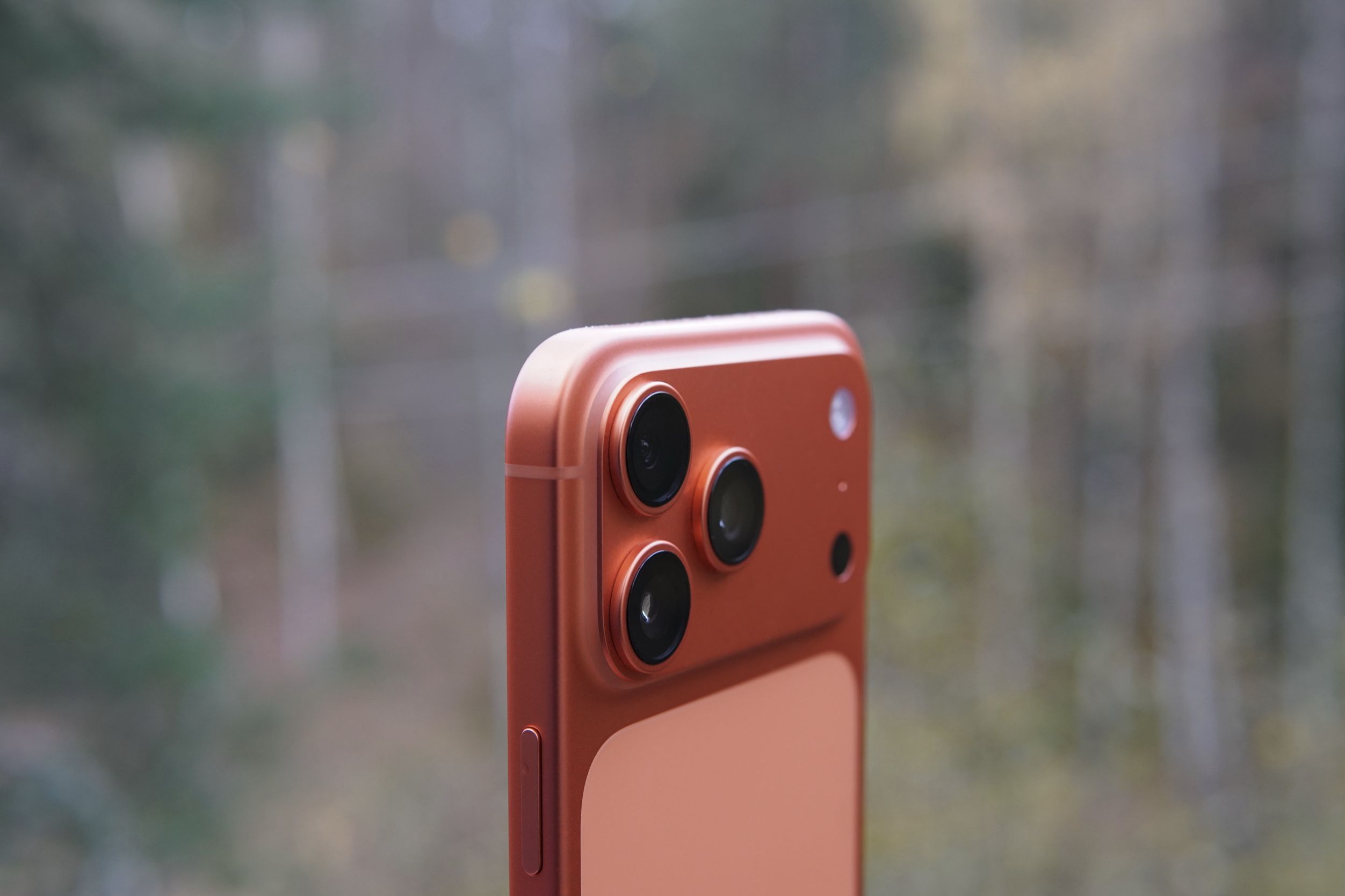 Close-up shot of a coral-colored smartphone, featuring three prominent camera lenses arranged in a triangular layout with a flash and small microphone hole, set against a blurred natural background.
