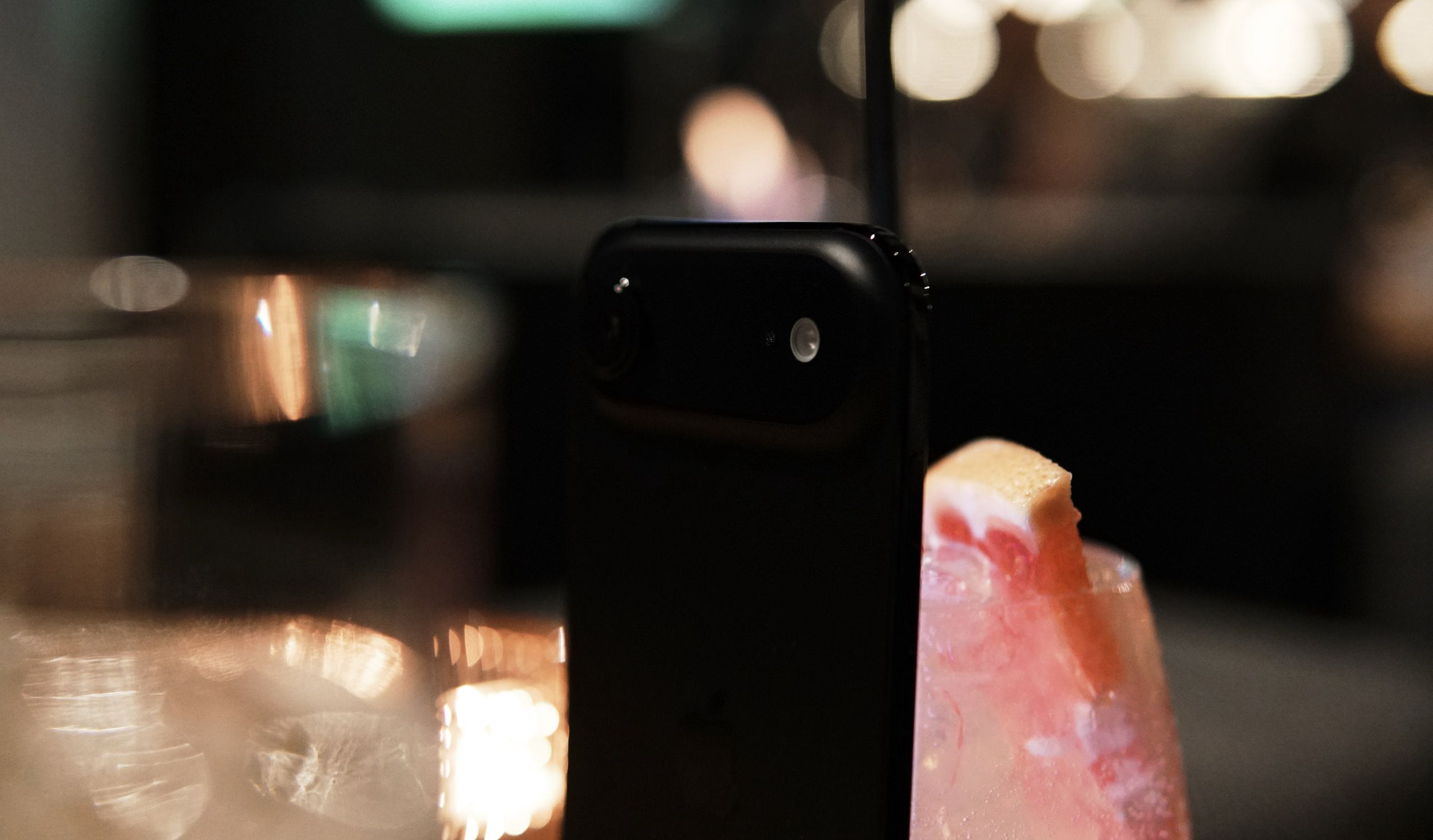 A partially obscured black smartphone rests near a cocktail garnished with a grapefruit wedge, with blurred background lights.
