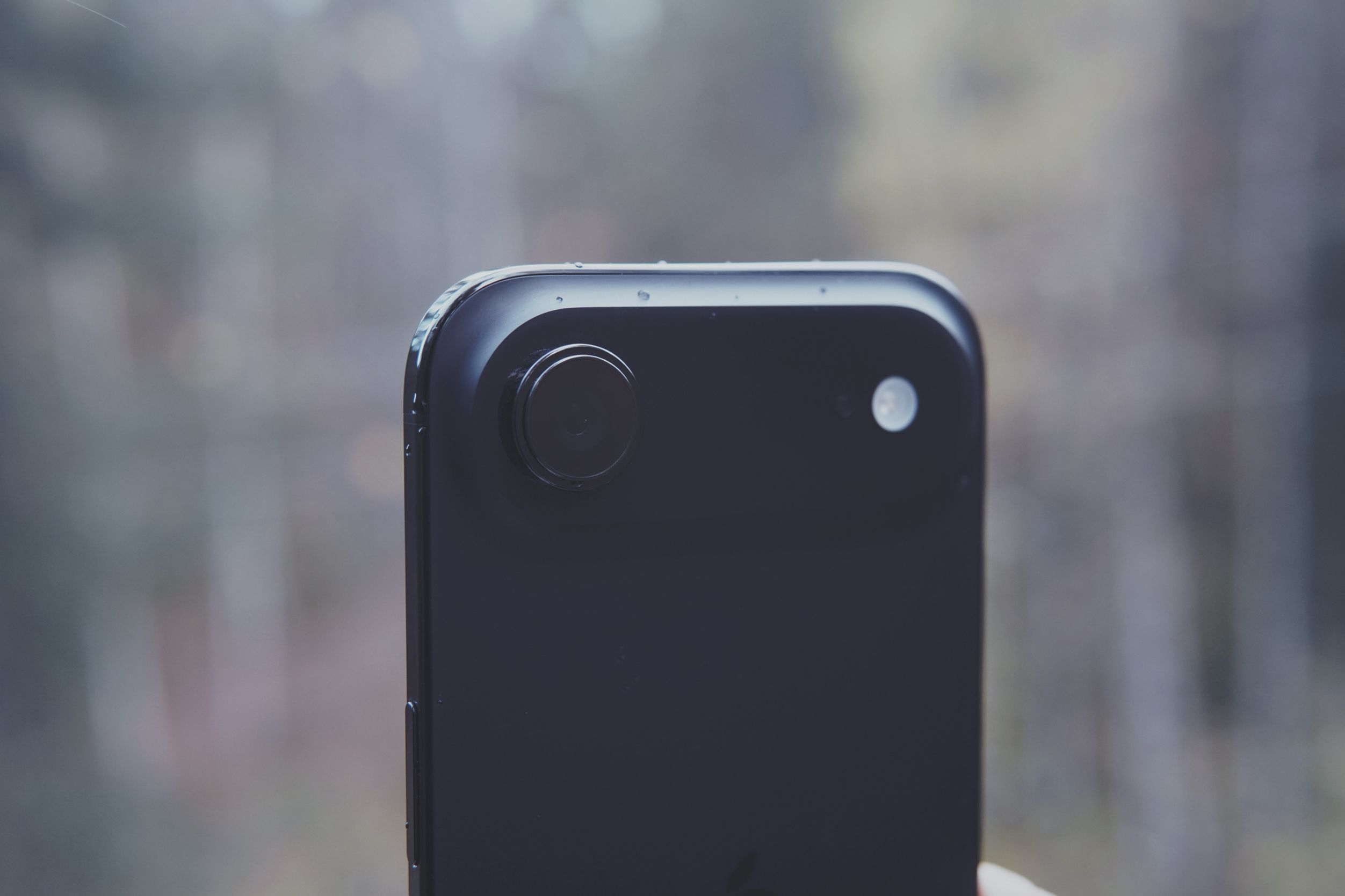 Close-up shot of the top portion of a black smartphone with a single, central, circular camera lens and an LED flash. The phone's surface has small water droplets. The background is blurred, suggesting an outdoor setting.
