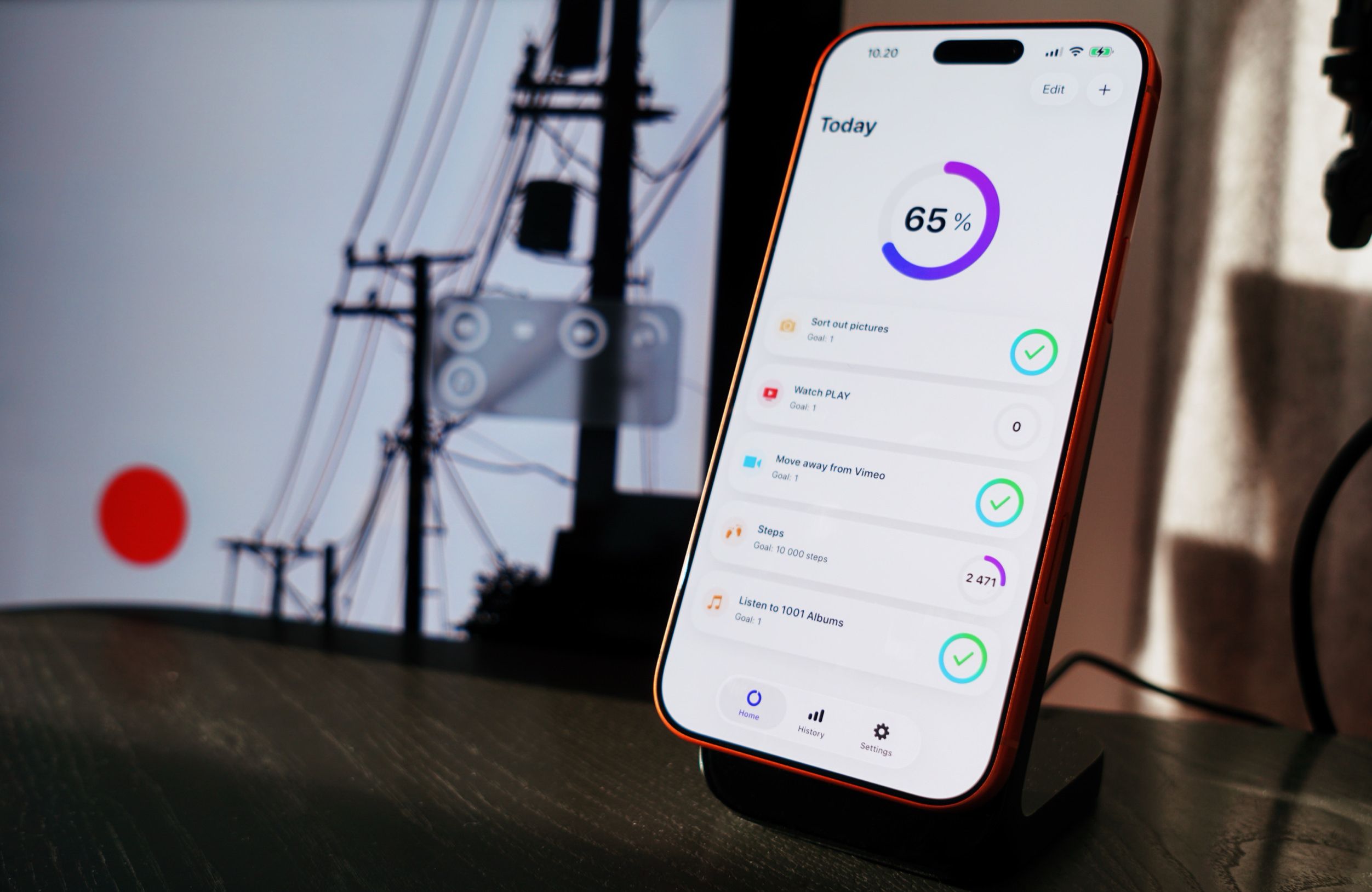 A smartphone displaying a productivity app, positioned on a dark desk with blurred out background elements; the phone is on a black stand showing today's progress as 65%, with tasks listed below.