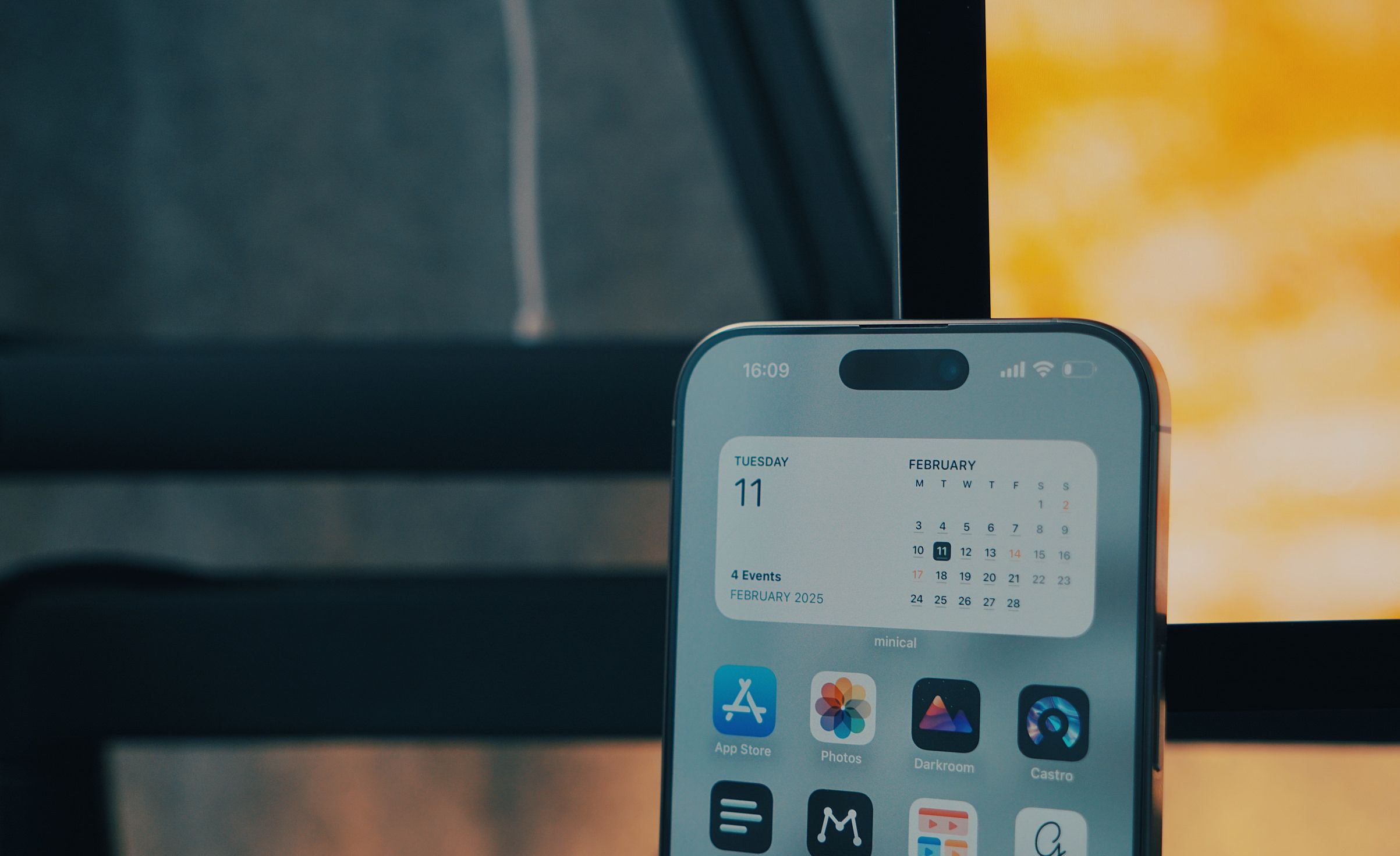 Close-up of a smartphone screen displaying the lock screen with the time "16:09" at the top, and a calendar widget. The widget shows "TUESDAY 11" and the month of "FEBRUARY". Below "4 Events FEBRUARY 2025." A calendar shows days of the month with the date "11" highlighted. Below the widget are app icons for "App Store", "Photos", "Darkroom", and "Castro", among others. The phone is in focus against a blurred background, suggesting a room or interior setting.
