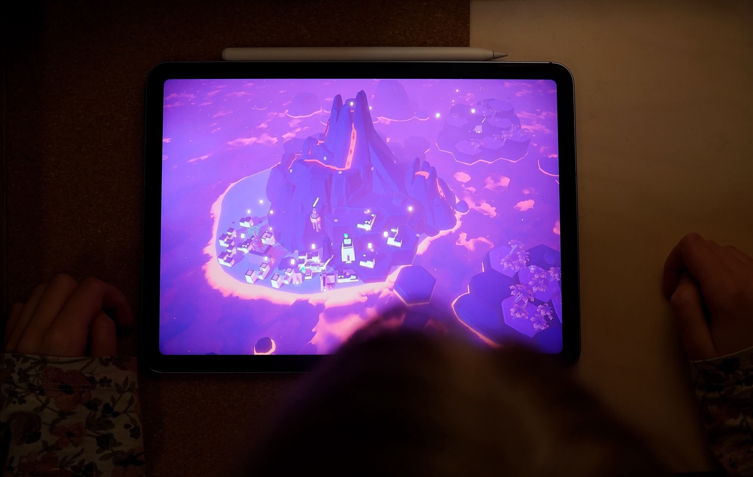 Eine Person, die ein Tablet hält, auf dem ein violett getöntes Fantasy-Videospiel mit schwebenden Inseln, Gebäuden und einer bergigen Struktur angezeigt wird.