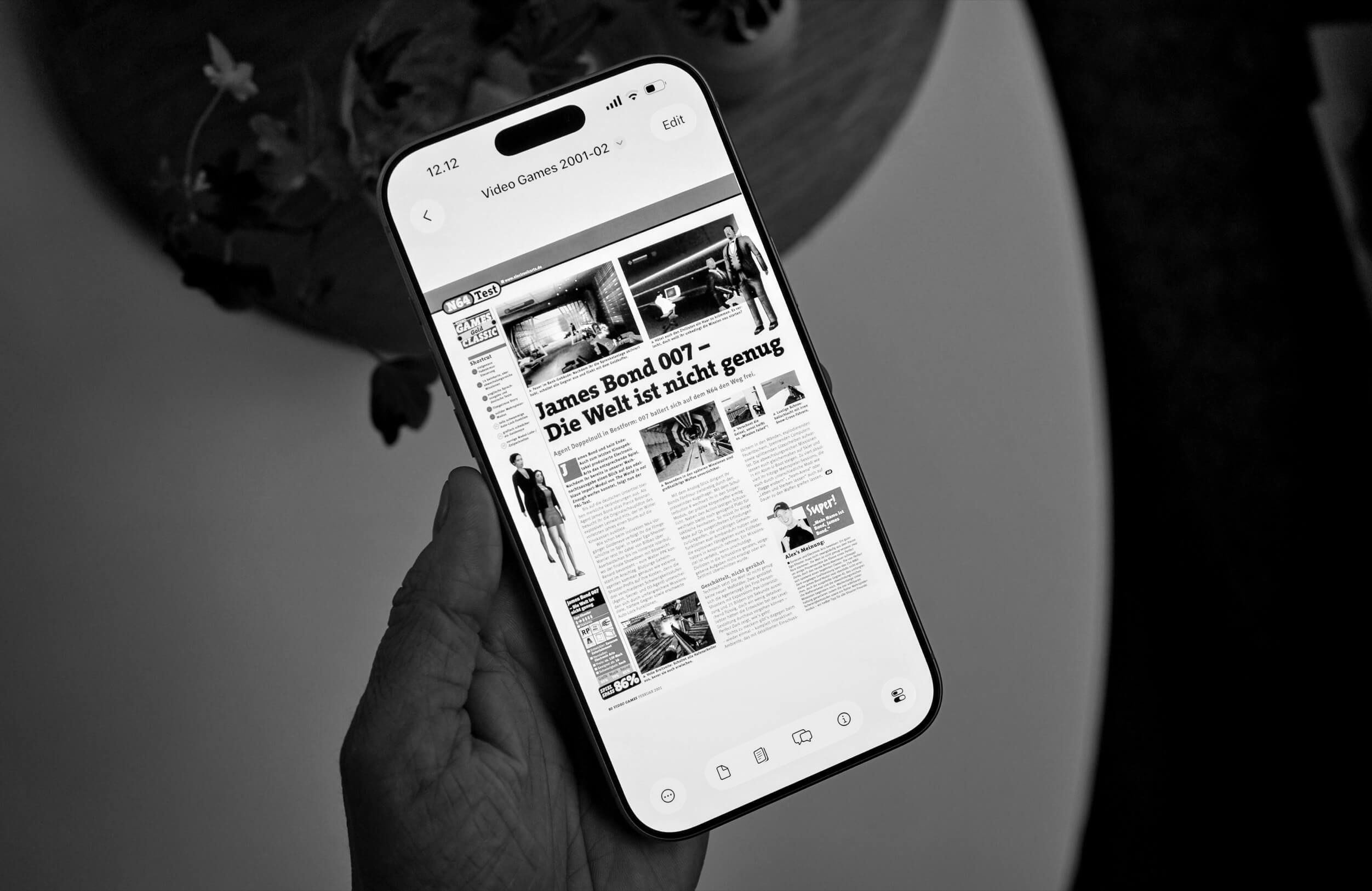 A hand holding a smartphone displaying a black and white image of a video game review. The review, in German, covers the James Bond 007 video game, featuring images from the game and the movie. The phone screen also shows the date and time, with an editing icon present.