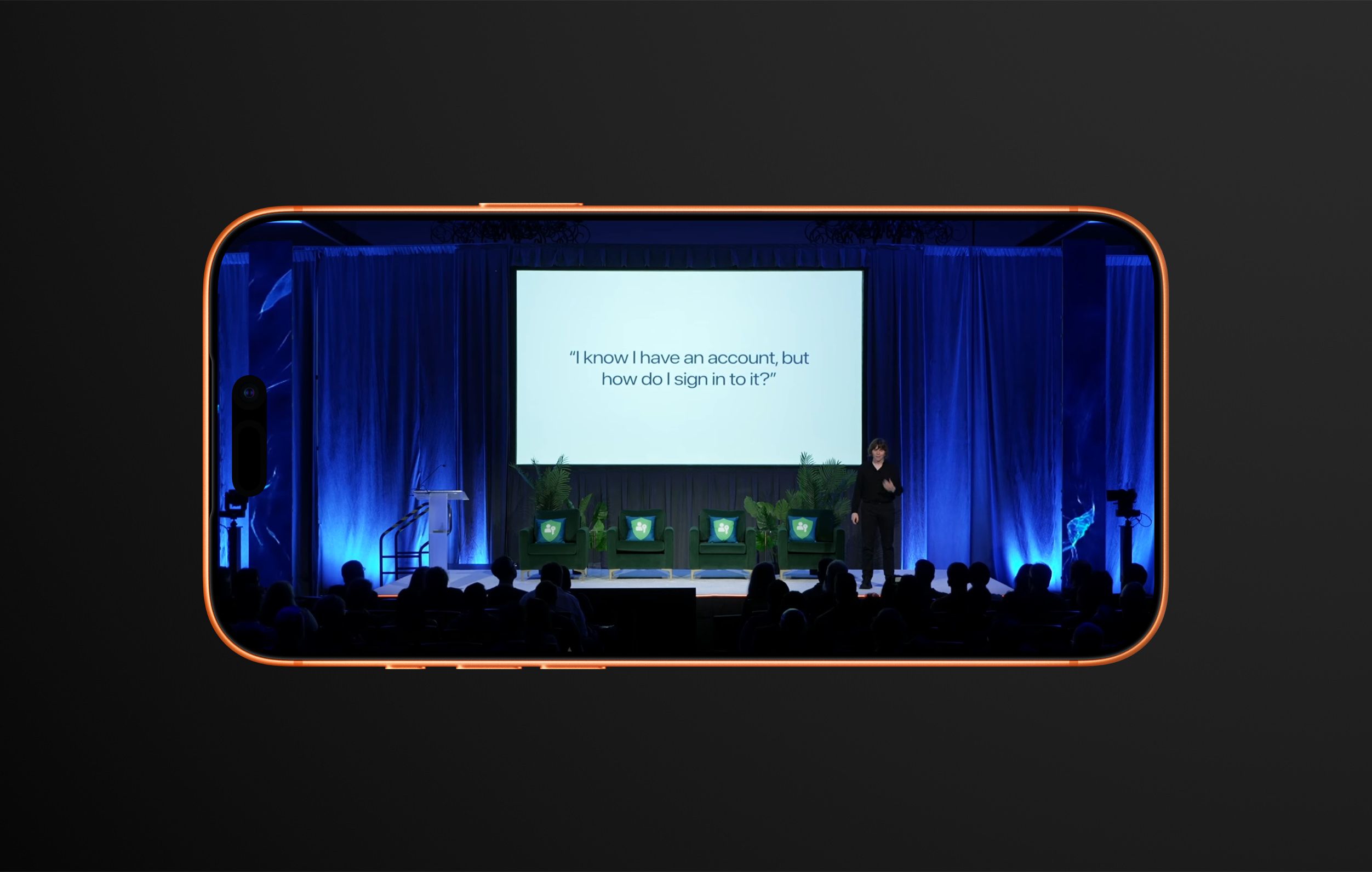 A presentation stage viewed through a stylized smartphone frame. A person in a black suit stands in front of a white screen displaying the text: "I know I have an account, but how do I sign in to it?". Behind the presenter are blue stage curtains, three green armchairs with throw pillows, and an audience in shadow. A podium and stage lighting are also visible. The phone frame is outlined in orange.