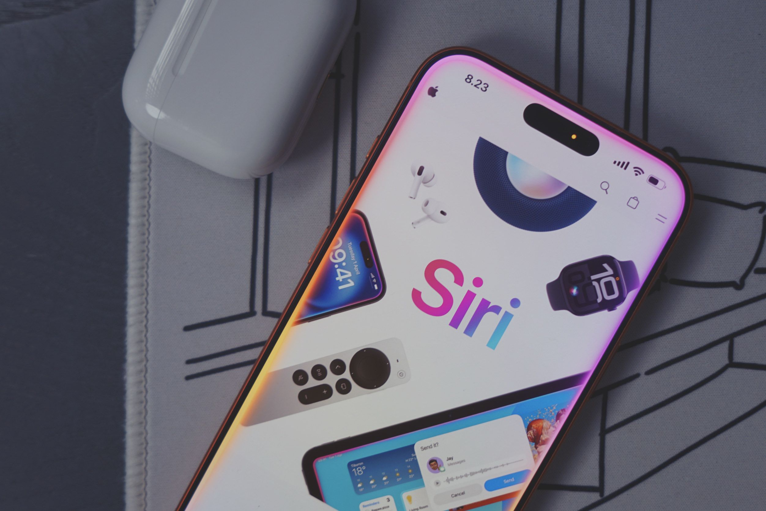 Close-up shot of an Apple iPhone displaying a Siri interface, various Apple products, and the time “8.23” in the top left corner of the screen. The screen is light-colored and features a large “Siri” logo. Surrounding the “Siri” logo, a pair of white Apple AirPods, a smartwatch with the number “10” on the screen, a remote control with a circular pad in the center, and a display with the temperature of “18°.” Additional screens on the iPhone display “Tuesday 1 April 09:41.” In the bottom of the screen, there is a display including “Send it?” and the name “Jay” with “Send” and “Cancel” buttons. A white Apple AirPod case rests beside the phone. The phone and AirPods case rest on a grey patterned surface.