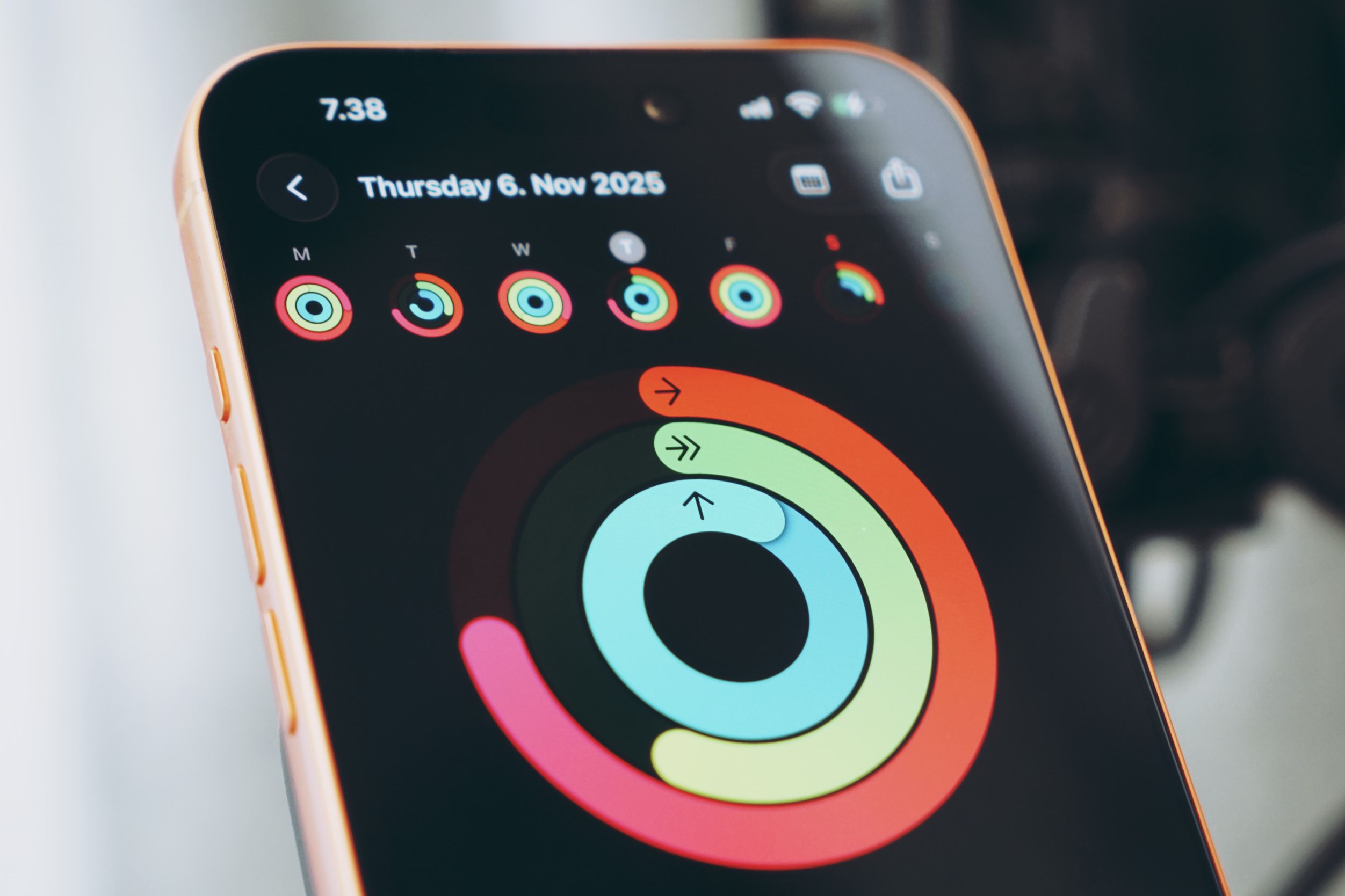 A close-up shot of a smartphone screen displaying an activity tracker app. The time at the top of the screen is "7.38" and the date "Thursday 6. Nov 2025" is shown below that. The app displays fitness rings, with three colored rings: pink, green, and blue, representing activity goals. The pink ring is partially filled, the green ring is mostly filled, and the blue ring is at least halfway filled. There is a "back" arrow at the top left of the screen and various notification icons at the top right. The phone has a gold-colored frame, and the background is blurred.