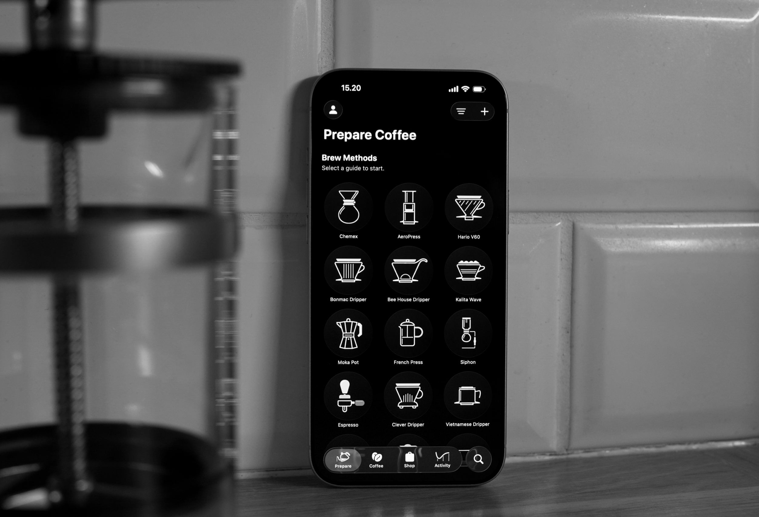 A grayscale image of a smartphone displaying a coffee brewing app, with a french press in the blurry foreground. The app's screen is black with white line icons and text. The app is in "Prepare" mode and displays a list of coffee brewing methods, including "Chemex", "AeroPress", "Hario V60", "Bonmac Dripper", "Bee House Dripper", "Kalita Wave", "Moka Pot", "French Press", "Siphon", "Espresso", "Clever Dripper", and "Vietnamese Dripper." The phone is set against a tiled wall.