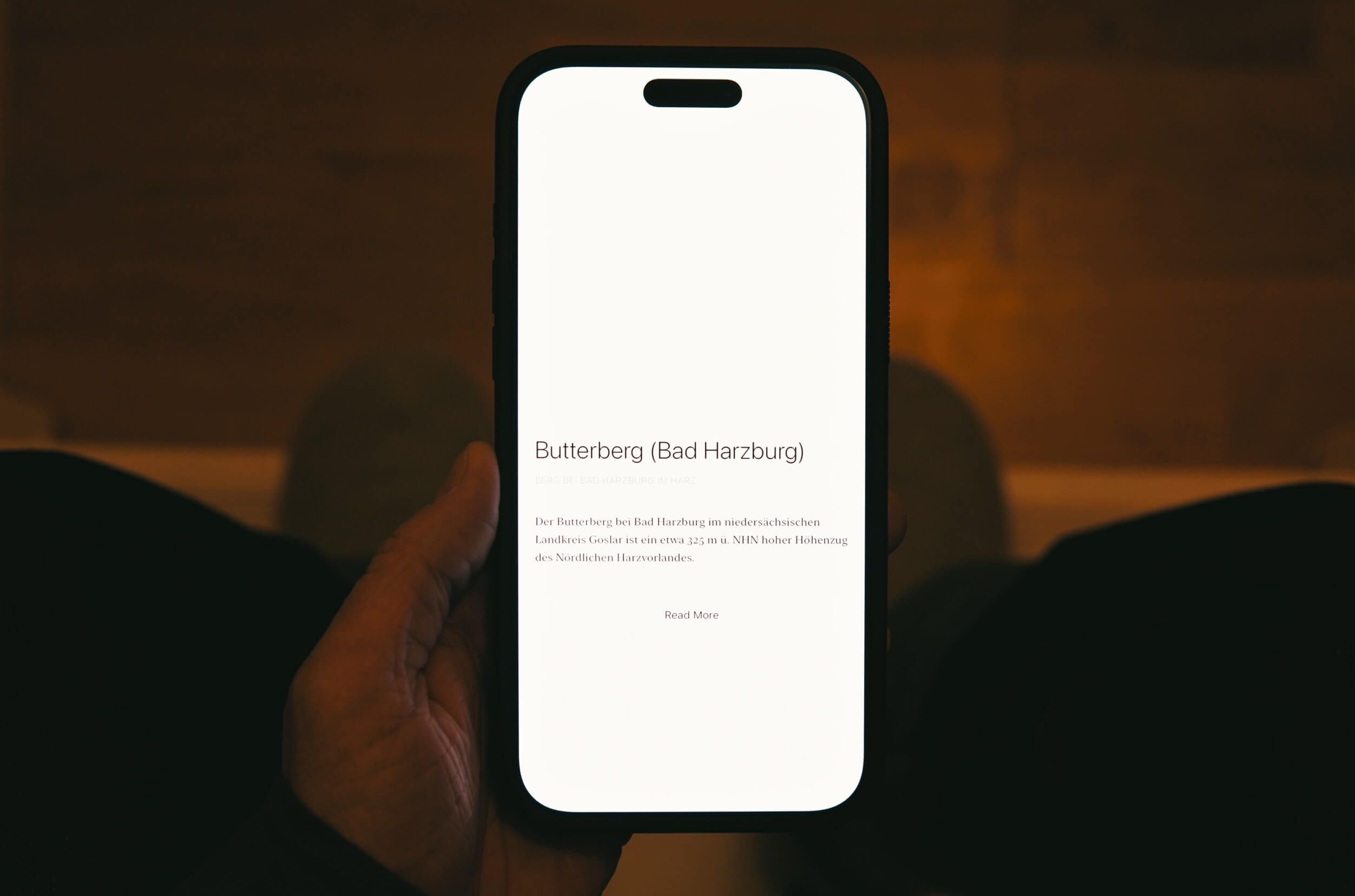 Eine Hand hält ein modernes Smartphone, dessen Bildschirm Informationen über den Butterberg (Bad Harzburg) auf Deutsch anzeigt, mit Text und einer Schaltfläche „Mehr lesen“; die Umgebung hat ein warmes, braunes Ambiente.
