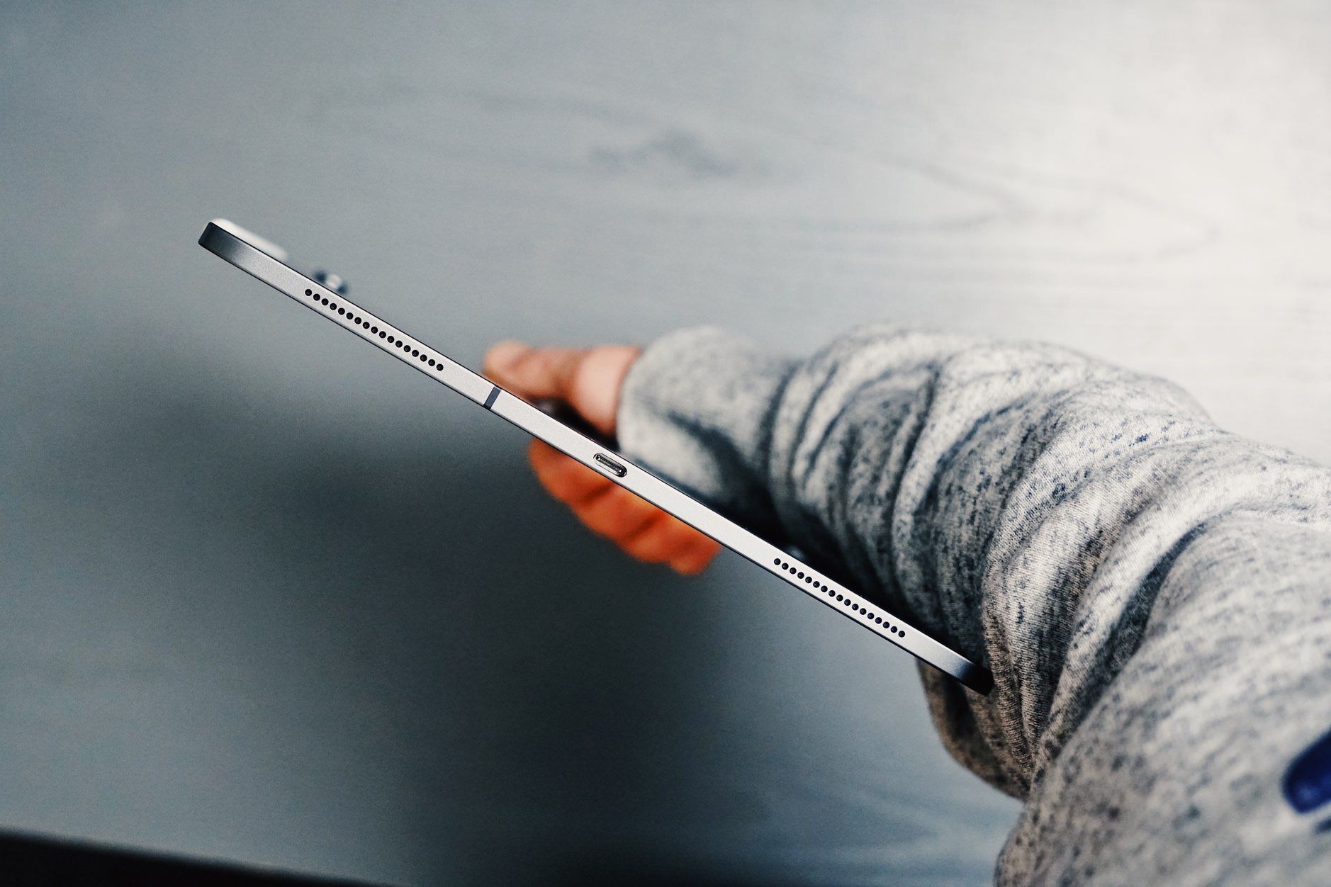 Hand hält ein silbernes Tablet und zeigt die Seite mit Lautsprecherlöchern und Ladeanschluss vor einem verschwommenen grauen Hintergrund, wobei ein grauer Hoodie getragen wird.
