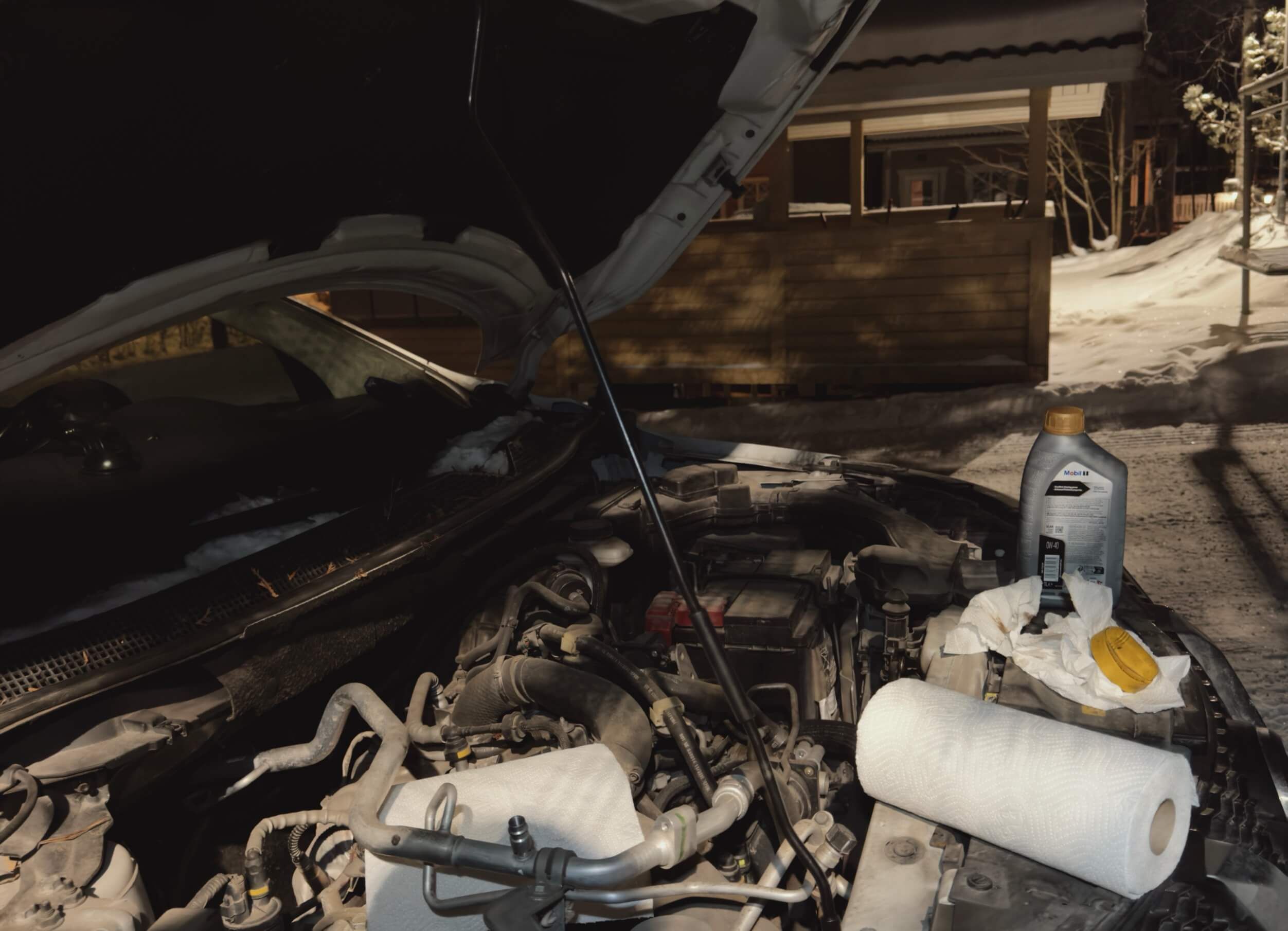 Motorraum eines Autos mit geöffneter Motorhaube, Papiertüchern, einer Ölflasche und einer gebrauchten Ölfilterkappe, vermutlich für einen Ölwechsel; verschneite Umgebung im Freien.