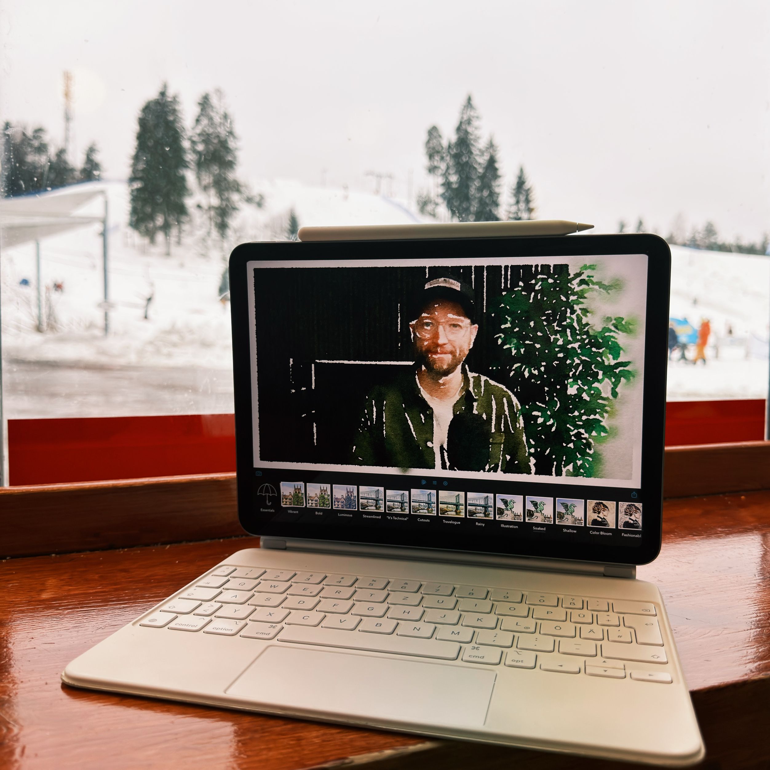 Ein Tablet mit abnehmbarer Tastatur zeigt einen Videoanruf eines Mannes mit Baseballkappe und Brille. Das Tablet befindet sich auf einer Holzoberfläche vor einem Fenster, durch das eine verschneite Landschaft sichtbar ist. Ein Eingabestift liegt auf der Oberkante des Tablets.