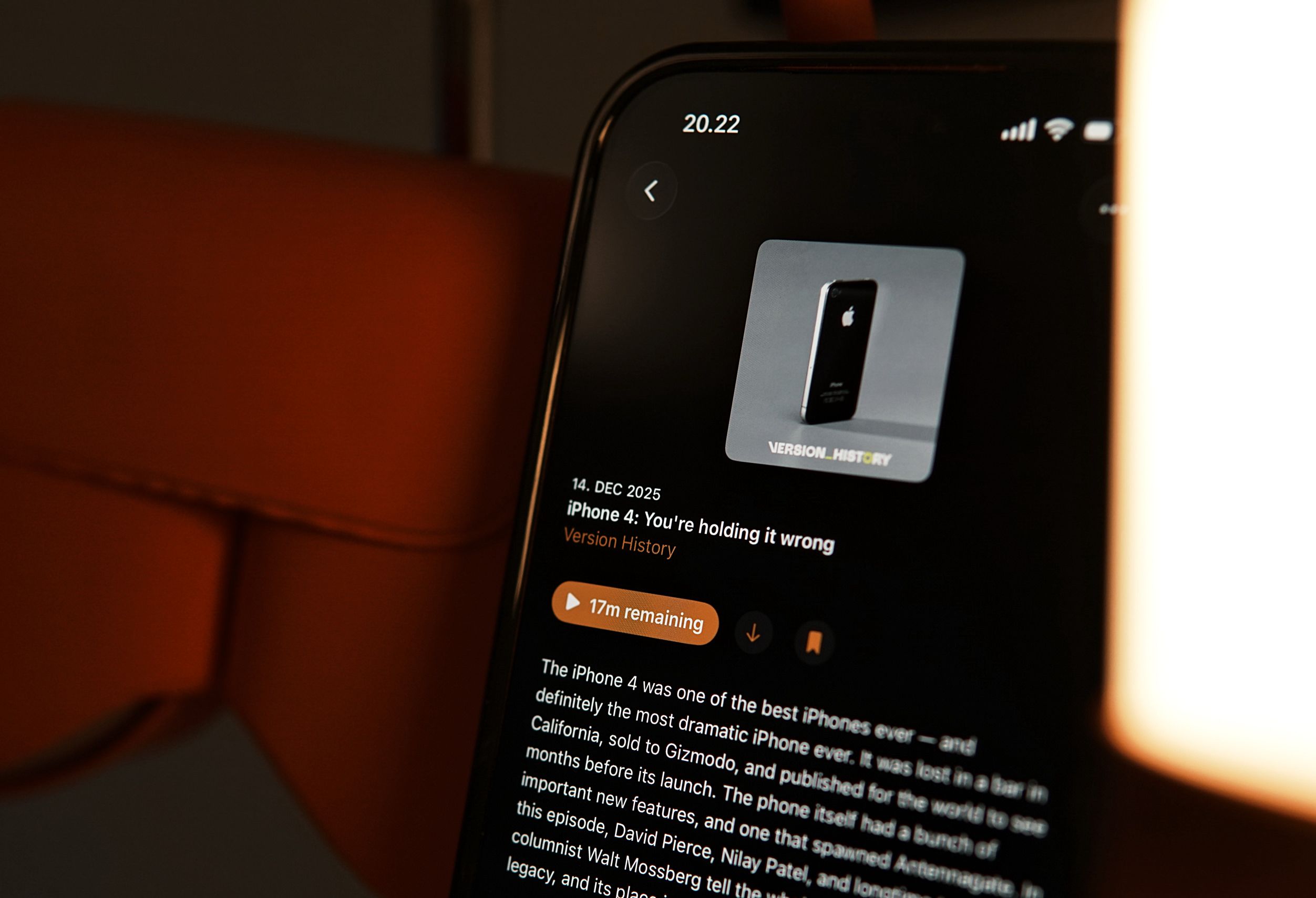 Nahaufnahme eines Smartphone-Bildschirms, der eine Podcast-Episode über das iPhone 4 anzeigt, mit einem Bild des Telefons und Episodendetails wie Titel, Datum und Restzeit. Das Telefon ist scharf gestellt, der Bildschirm leuchtet und der Hintergrund ist in warmen Orangetönen unscharf.