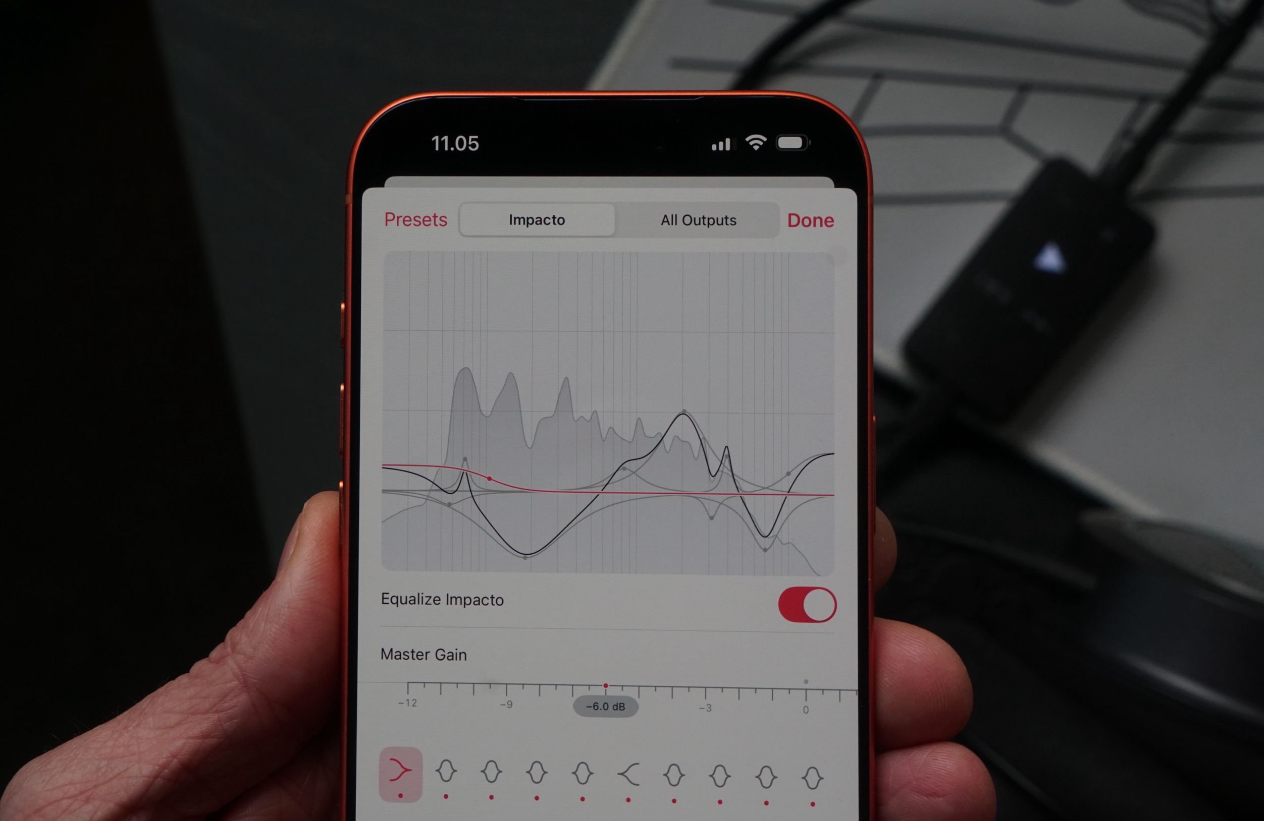 Eine Hand, die ein rotes Telefon hält, das eine Audio-Equalizer-Oberfläche anzeigt. Die Oberfläche zeigt ein Diagramm mit mehreren Linien und mehreren Optionen, darunter: "Presets", "Impacto", "All Outputs", "Done", "Equalize Impacto" und "Master Gain". Der Text auf dem Telefon lautet: "11.05".
