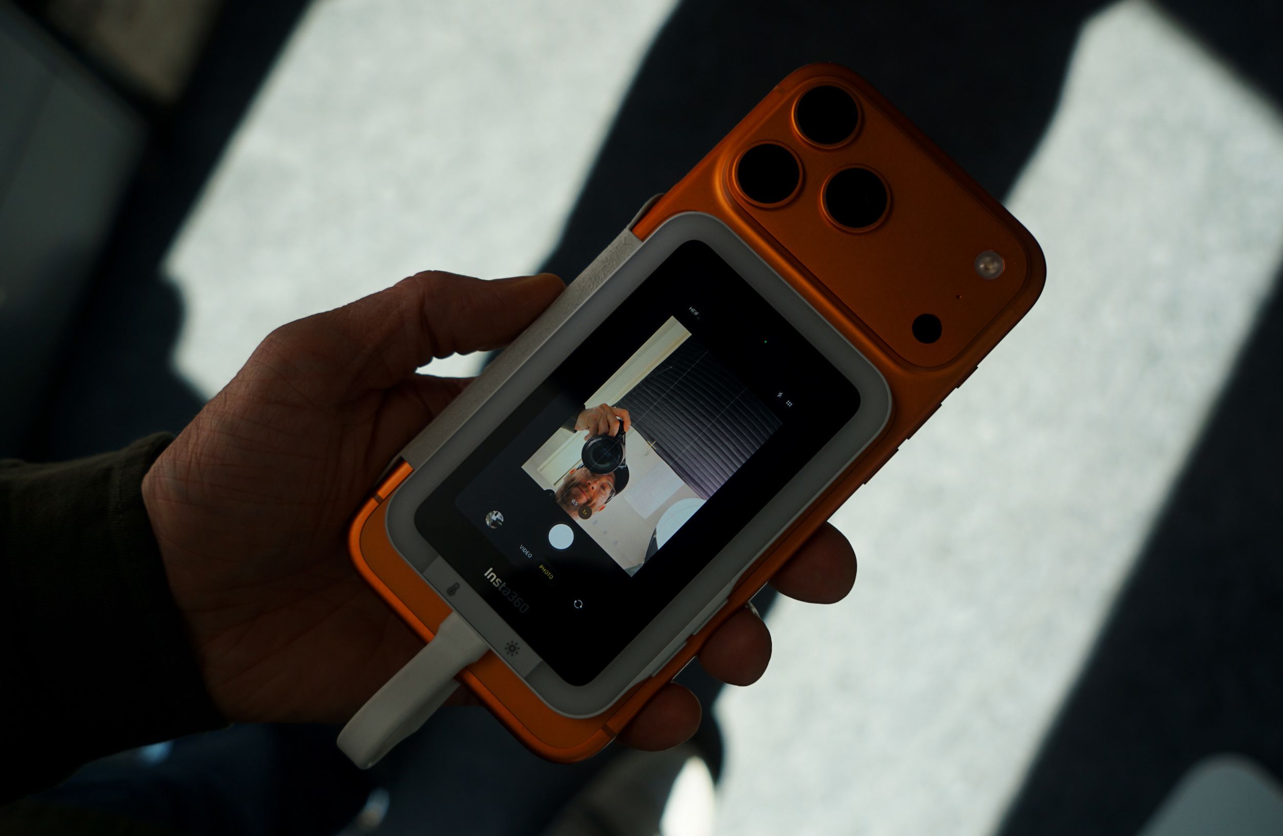 Eine Hand hält ein orangefarbenes Kamerazubehör, das an einem Smartphone befestigt ist. Auf dem Bildschirm ist eine Live-Kameraansicht zu sehen, die die Person beim Aufnehmen des Bildes zeigt. Am unteren Bildschirmrand steht der Text „Insta360“.