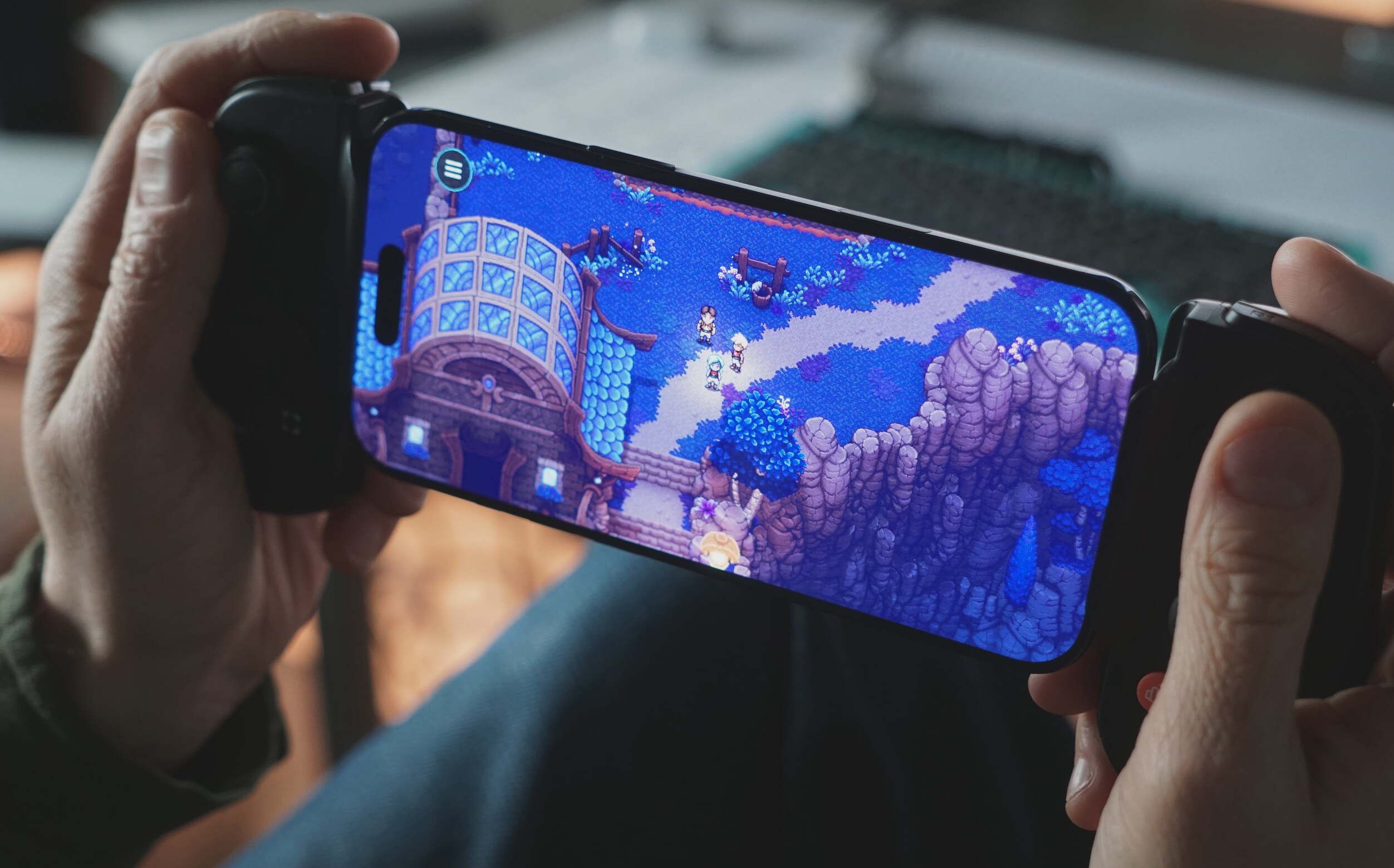 Eine Person hält ein Smartphone im Querformat mit einem beidseitig angebrachten mobilen Gaming-Controller. Auf dem Bildschirm ist ein Rollenspiel im Pixel-Art-Stil mit einem Gebäude, einem Pfad und kleinen Charakteren zu sehen. In der oberen linken Ecke befindet sich ein Menüsymbol.