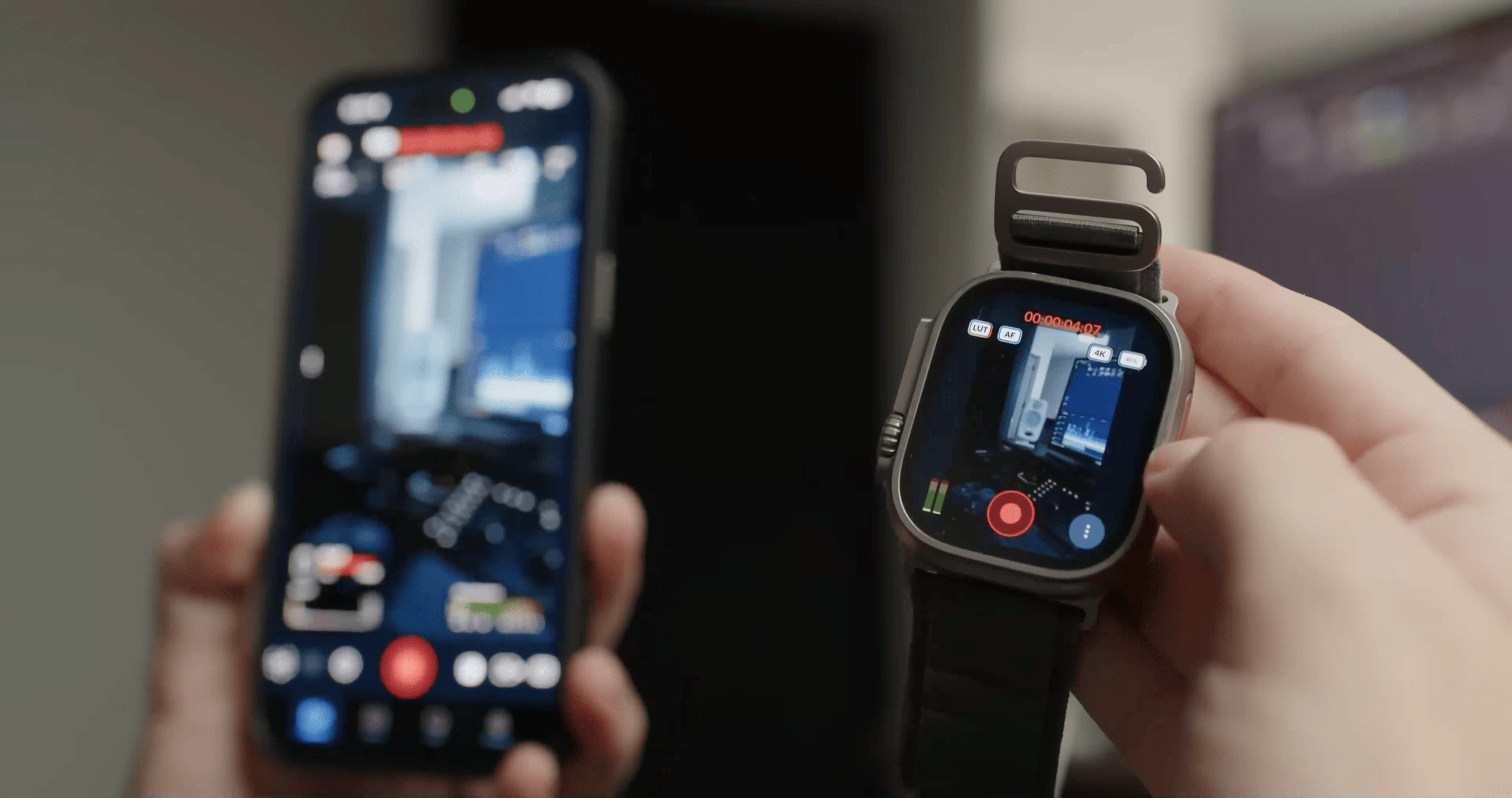 Eine Person hält ein Smartphone in der linken Hand und eine Smartwatch in der rechten Hand. Beide Bildschirme zeigen denselben Kamera-Sucher mit einer Ansicht eines Raumes.
