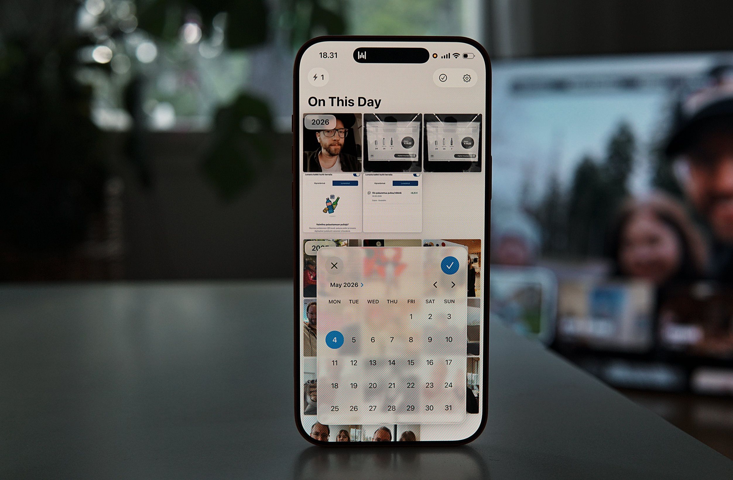 Ein Smartphone zeigt eine Fotogalerie-App mit der Überschrift „On This Day“. Ein halbtransparentes Kalenderfenster ist geöffnet, das den „Mai 2026“ anzeigt, wobei der 4. Tag blau markiert ist. Im Hintergrund sind unscharfe Bildvorschauen zu sehen.