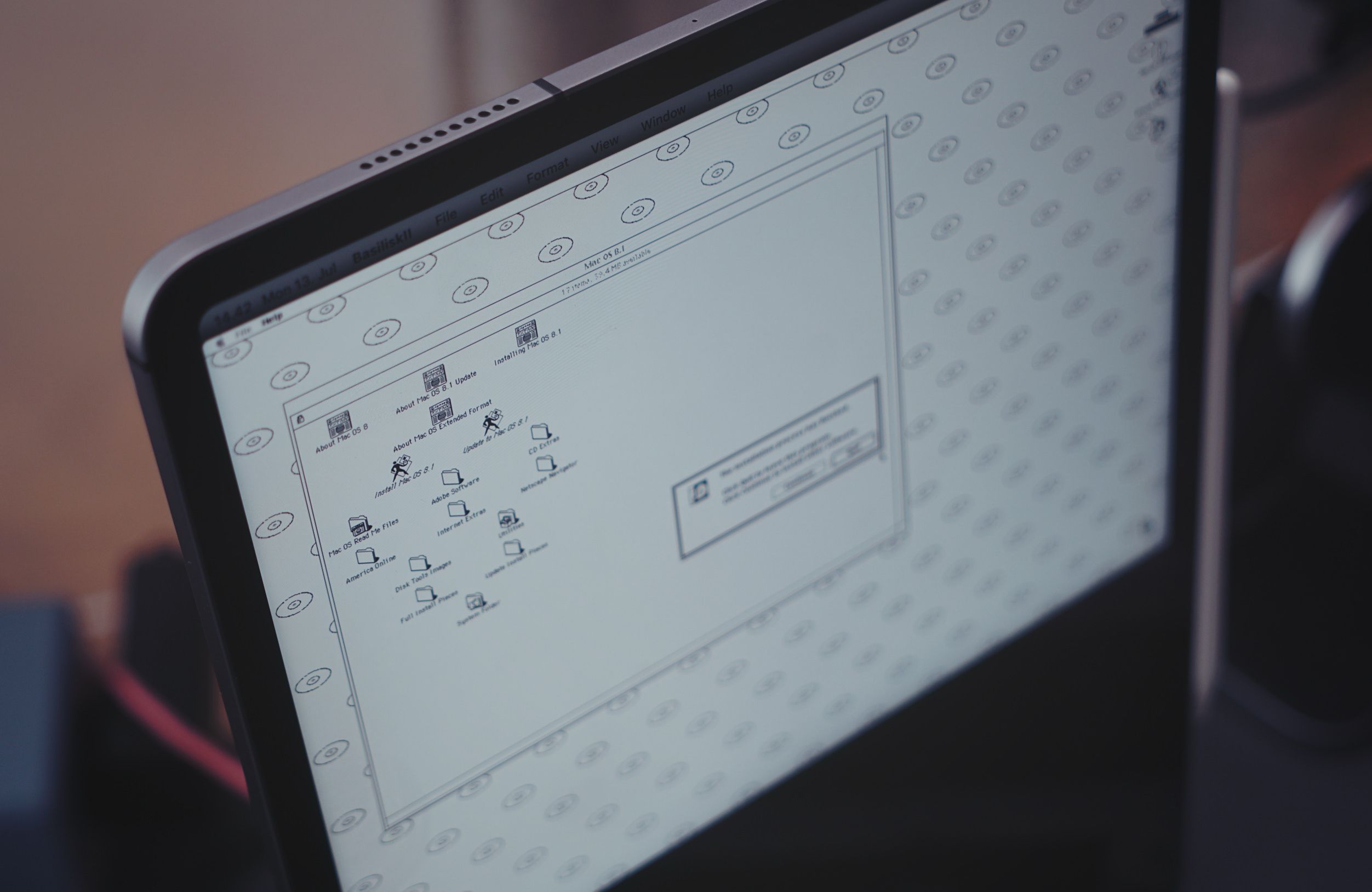 Ein Computerbildschirm zeigt eine klassische Mac OS-Desktop-Oberfläche mit verschiedenen Symbolen, Ordnern und einem geöffneten Fenster vor einem gemusterten Hintergrund.