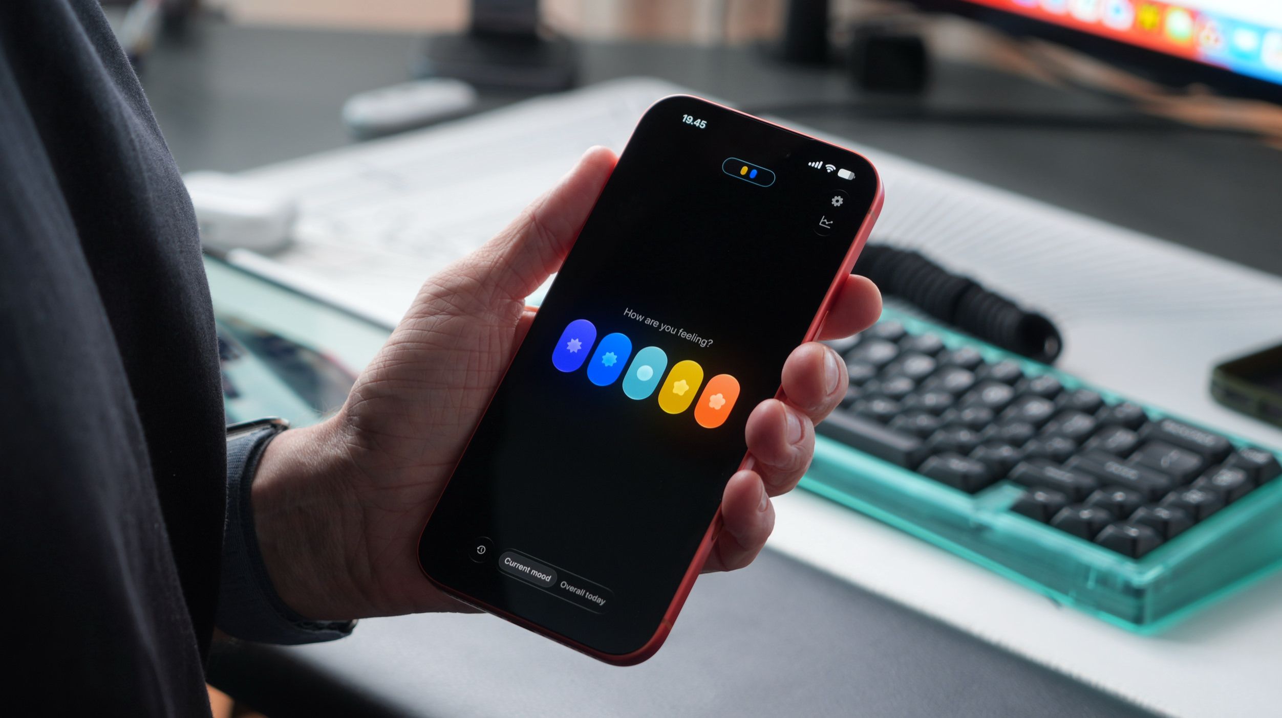Eine Person hält ein Smartphone, auf dessen Display die Benutzeroberfläche einer Stimmungs-App mit fünf bunten, blumenförmigen Symbolen unter der Frage „Wie fühlst du dich?“ in einer Büroumgebung angezeigt wird.