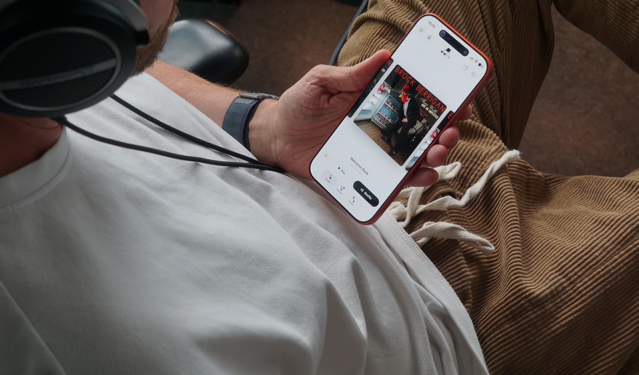 Eine Person in einem weißen T-Shirt, einer beigen Cordhose und Kopfhörern hält ein Smartphone mit einer Musik-App-Oberfläche in der Hand.