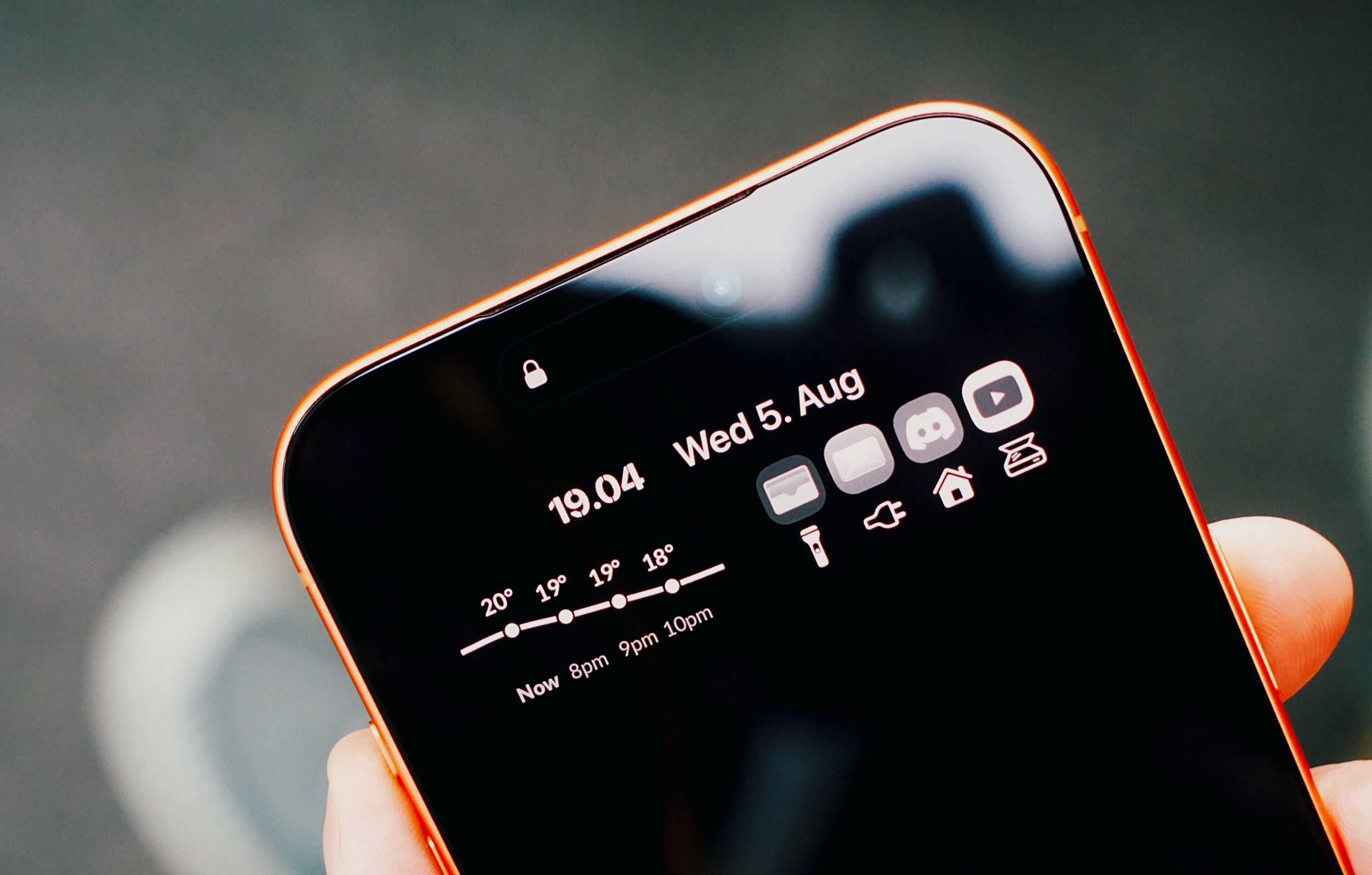 Nahaufnahme eines orangefarbenen Smartphones in einer Hand. Der Sperrbildschirm im dunklen Design zeigt die Uhrzeit, das Datum, ein Wettervorhersage-Diagramm sowie eine Anordnung verschiedener App-Symbole.