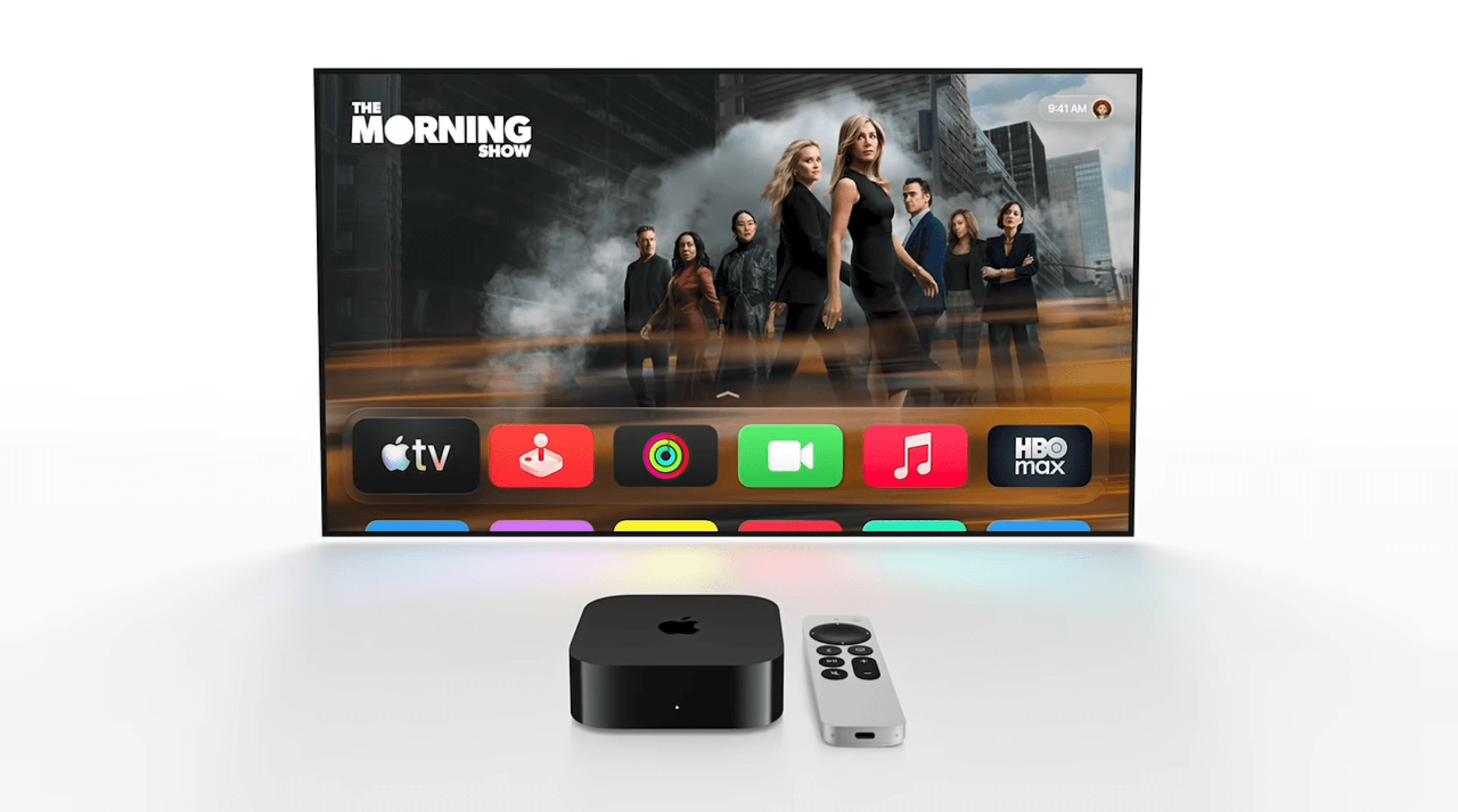 A flat-screen television displaying the show 'The Morning Show' with a row of app icons including Apple TV, a red joystick icon, activity rings, video call, music, and HBO Max at the bottom. In front of the television sits a black Apple TV box and a silver remote control on a white surface.