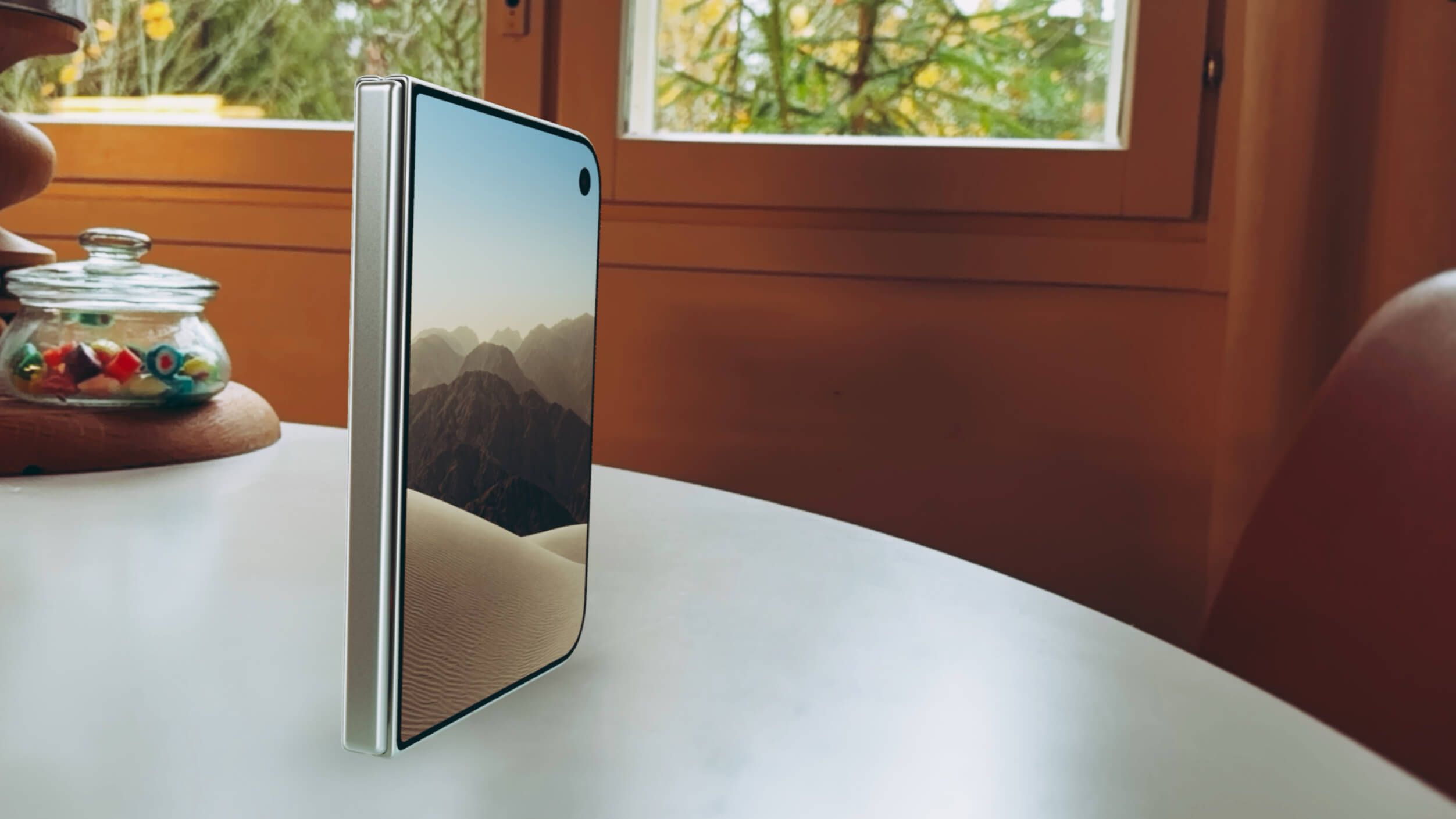 A modern foldable smartphone in a partially folded state standing upright on a round white table. The screen displays a desert landscape with mountains and a clear sky.