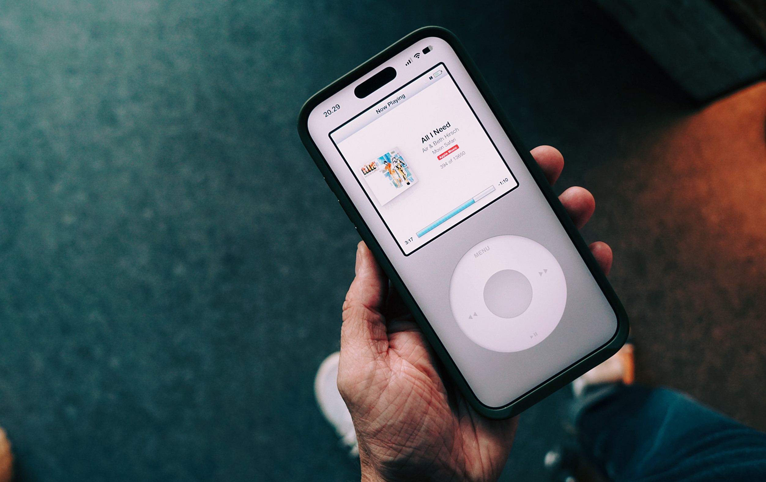 Eine Nahaufnahme einer Hand, die ein Smartphone hält, auf dessen Display eine Musik-App im Stil eines klassischen iPods mit Click-Wheel angezeigt wird.