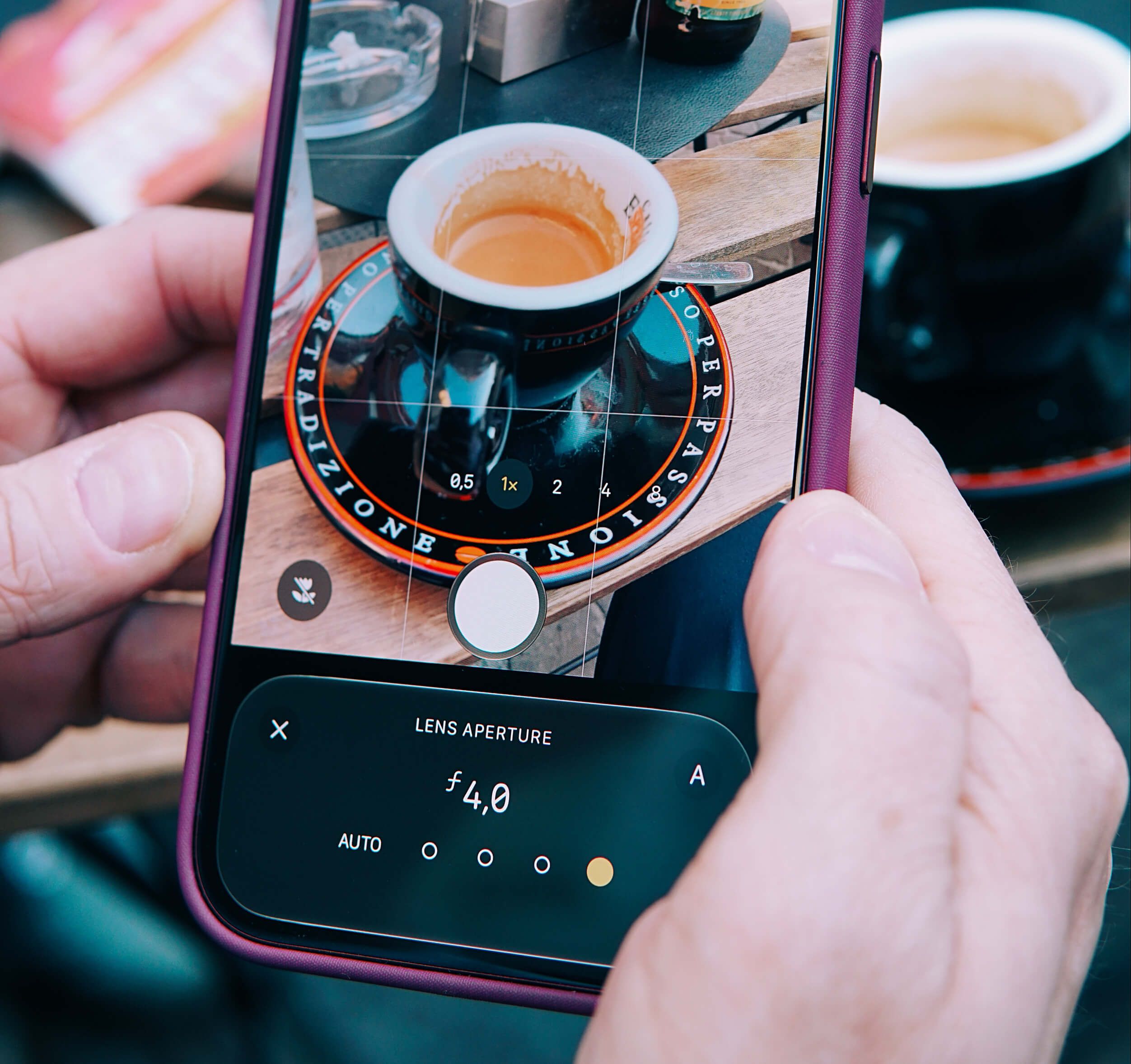 A close-up of a person holding a smartphone taking a photo of a small coffee cup on a saucer placed on a wooden table. The phone screen shows camera settings including a lens aperture of f4.0, and the focus is on the coffee cup in the center of the frame.